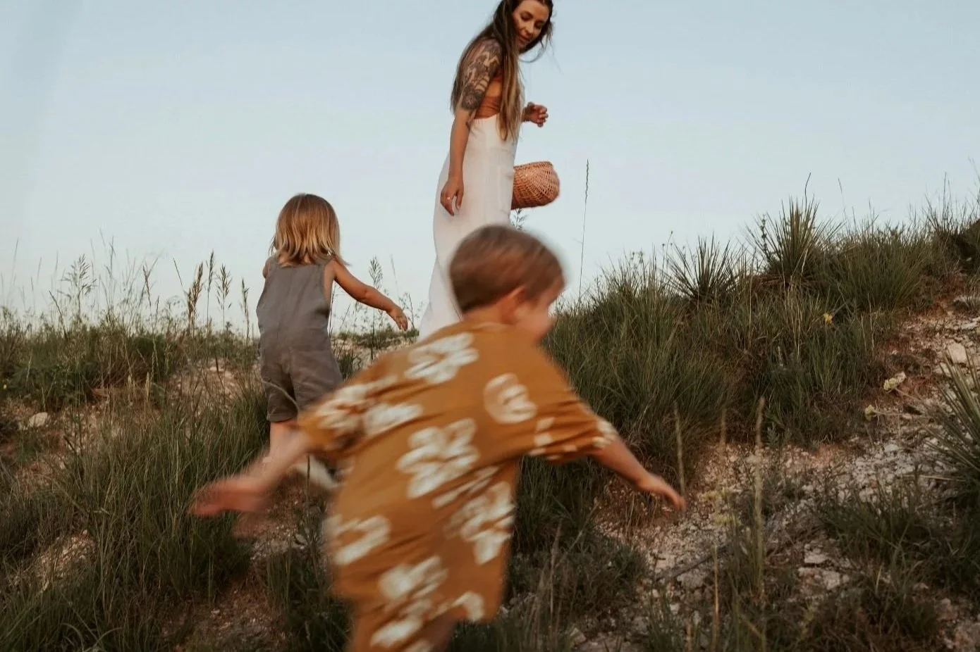 A woman with long hair and tattoos, carrying a basket, guides two young children in casual summer clothing up a grassy hill outdoors during sunset.