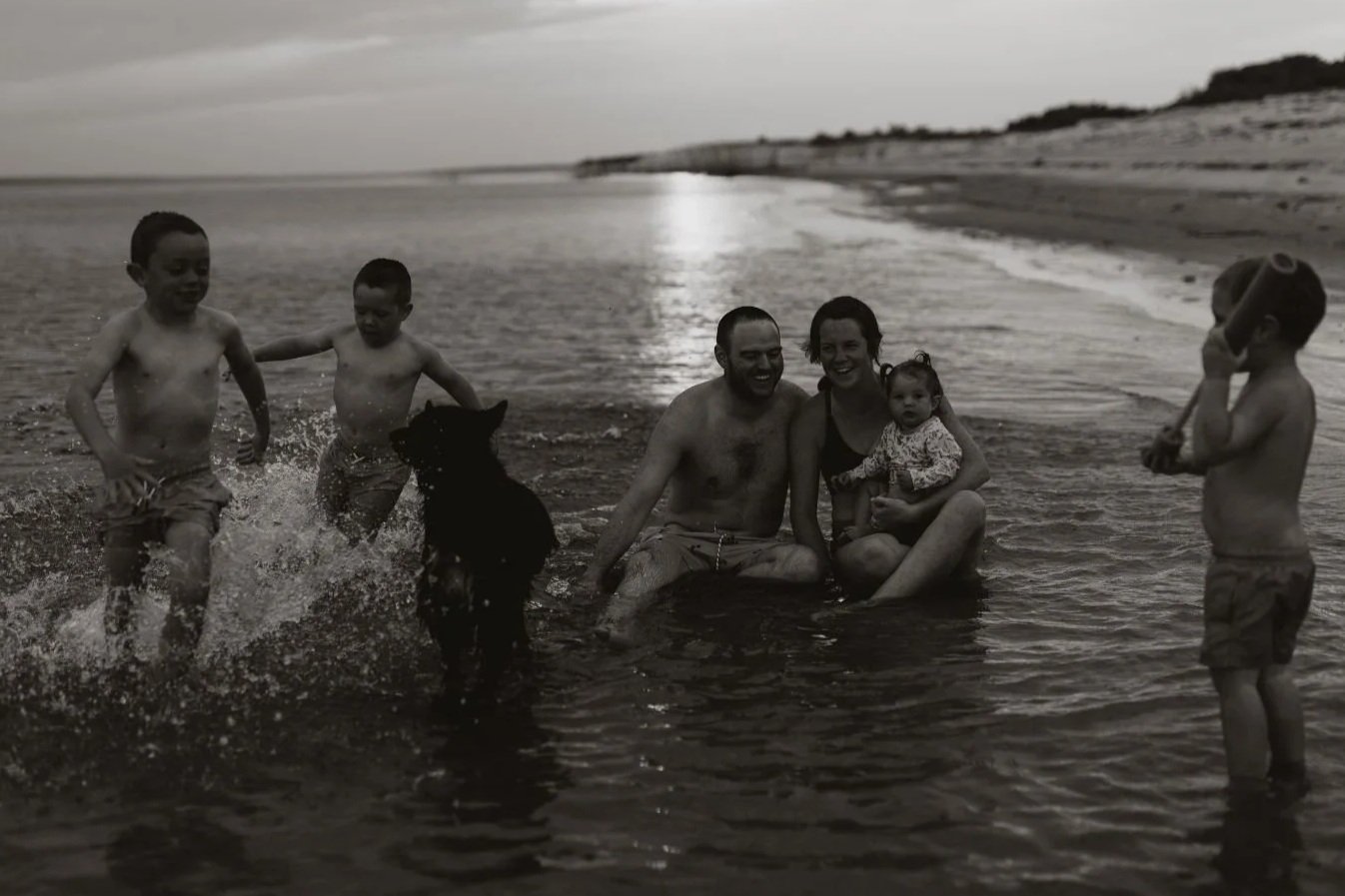 Family enjoying time in the ocean with children playing and an adult couple sitting in water, a dog jumping out of the water, and a young child talking on a toy phone.