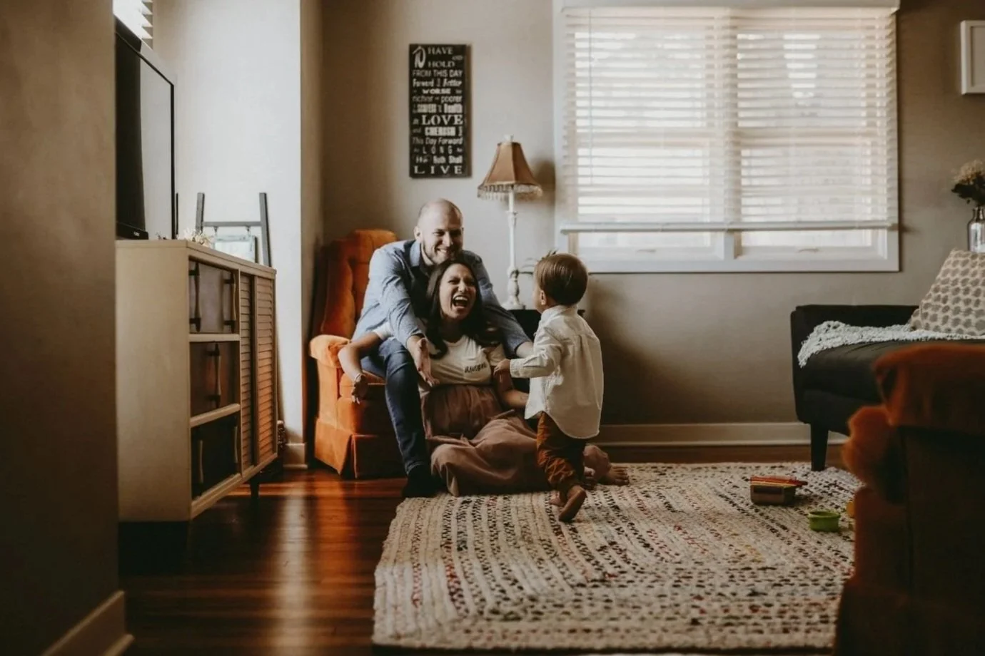 Family playing and laughing together in living room, with a woman sitting on the floor, a man sitting behind her, and a young boy standing in front, holding her hands.