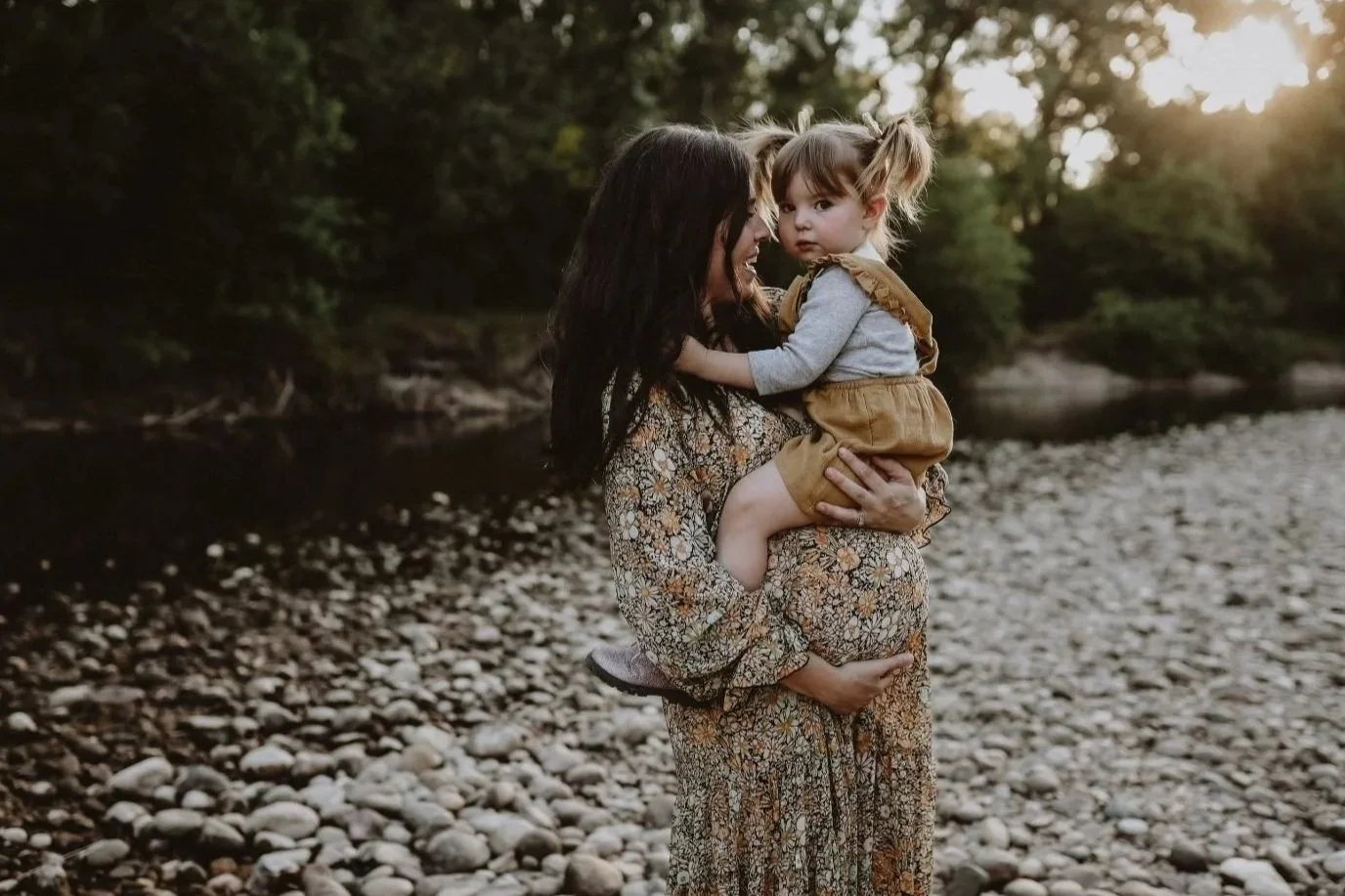 A woman with long dark hair wearing a floral dress holds a young girl with pigtails and light hair, dressed in a gray top and tan overalls, on a rocky riverside during sunset.