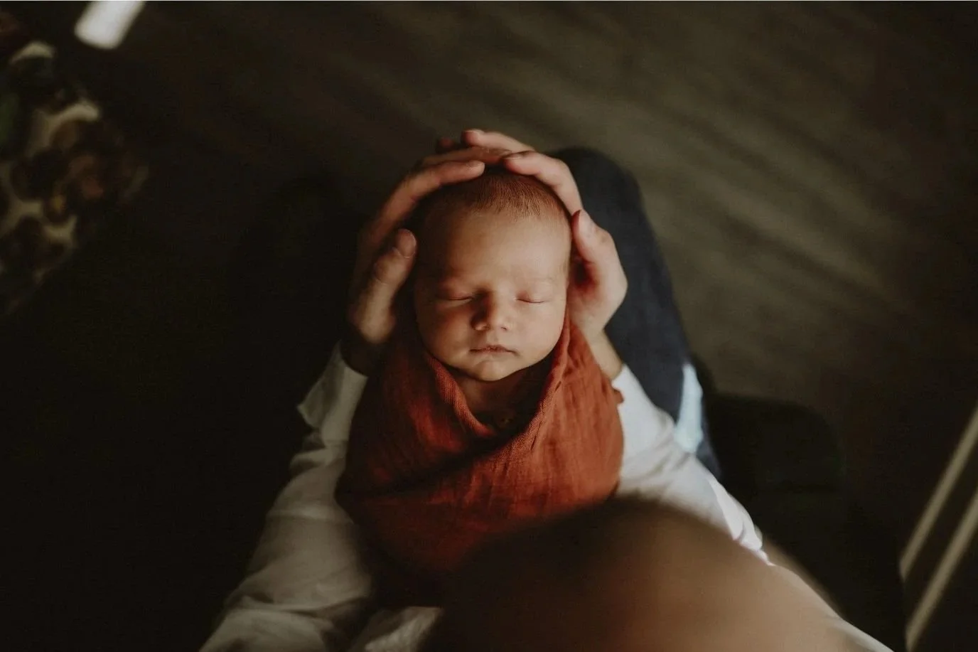 A baby with closed eyes being gently held on the head and shoulders by an adult, resting on the adult's lap. The baby is wrapped in a reddish-brown blanket and wearing a white outfit, with a wood-textured background.