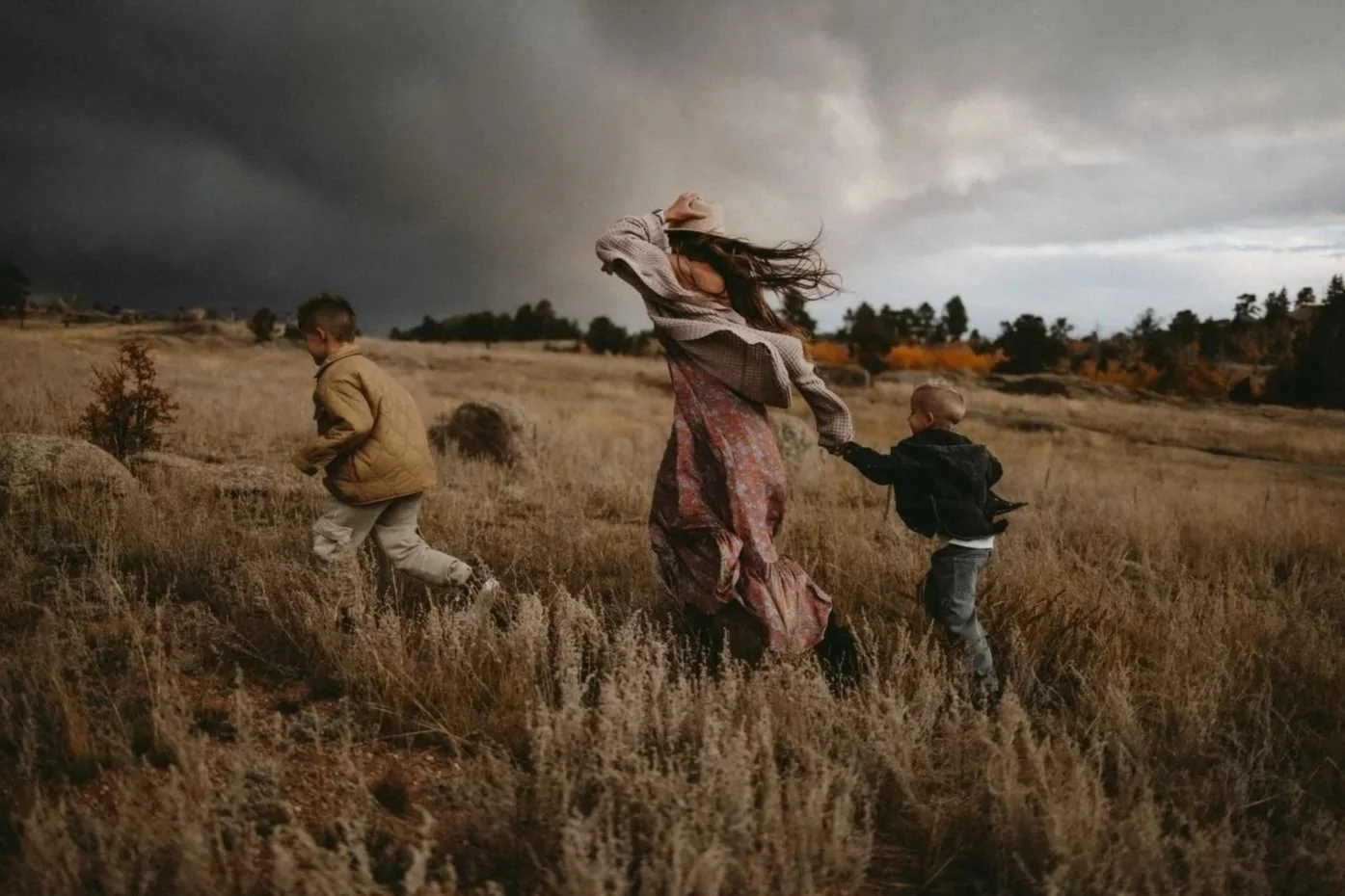 A woman with long hair and a floral skirt running through a field with two children, holding hands with one, as dark storm clouds loom overhead.