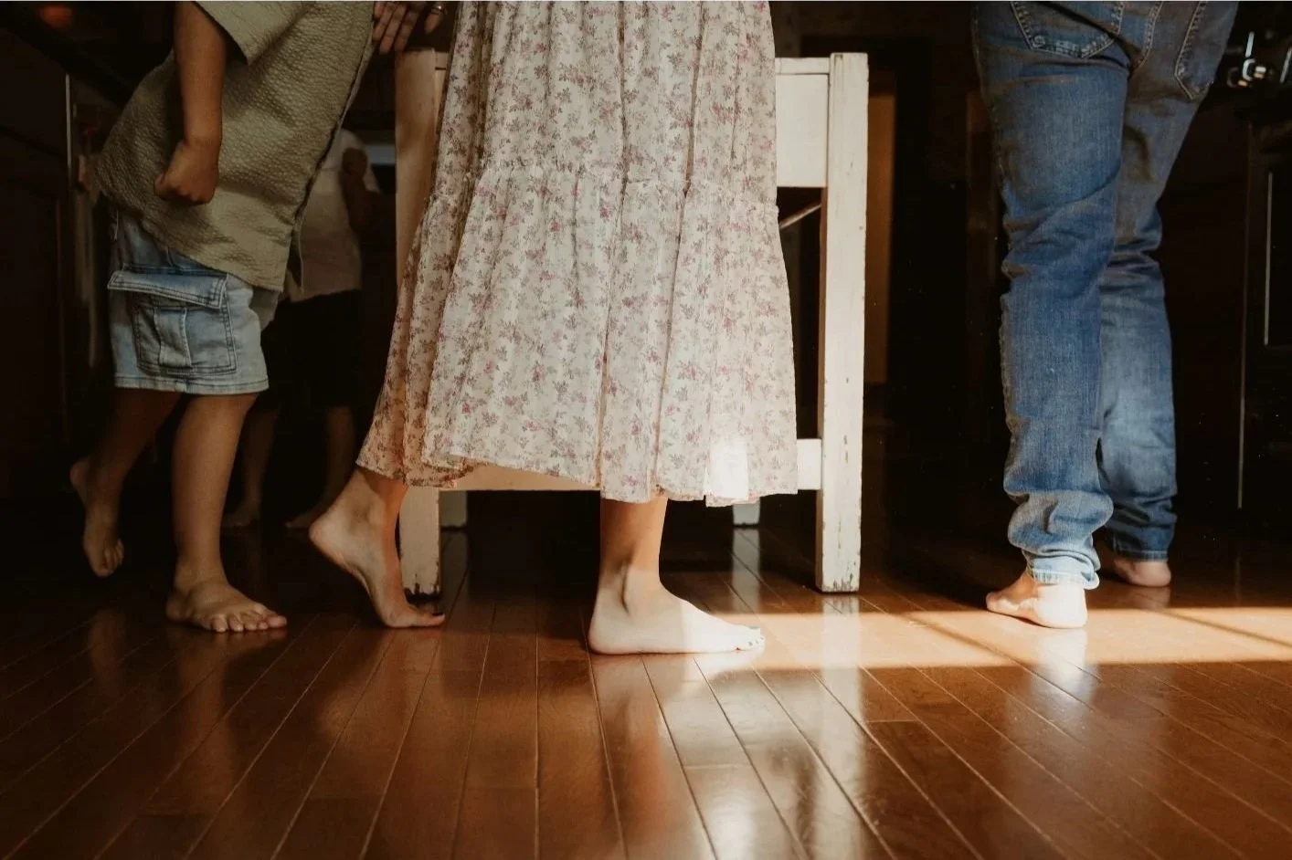 People standing barefoot on a wooden floor in a dimly lit room, with clothing indicating it is a casual setting, and a white wooden table in the background.