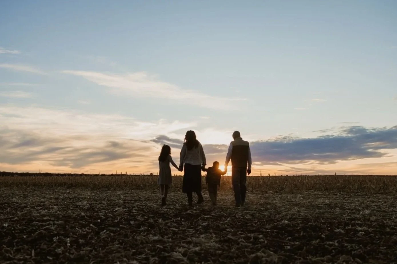 A family of four holding hands walking in a field at sunset, with the sun on the horizon and a partly cloudy sky.
