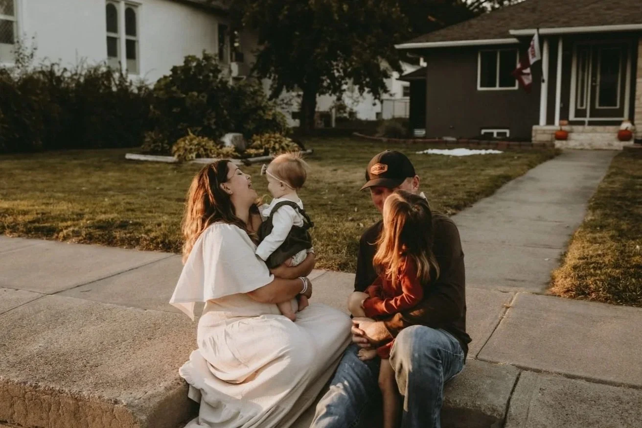 A family enjoying a happy moment outdoors on a sidewalk in the evening. The woman is sitting on the curb, holding a baby girl and smiling at her. A young girl with brown hair is sitting next to her, and a man wearing a baseball cap is sitting beside 