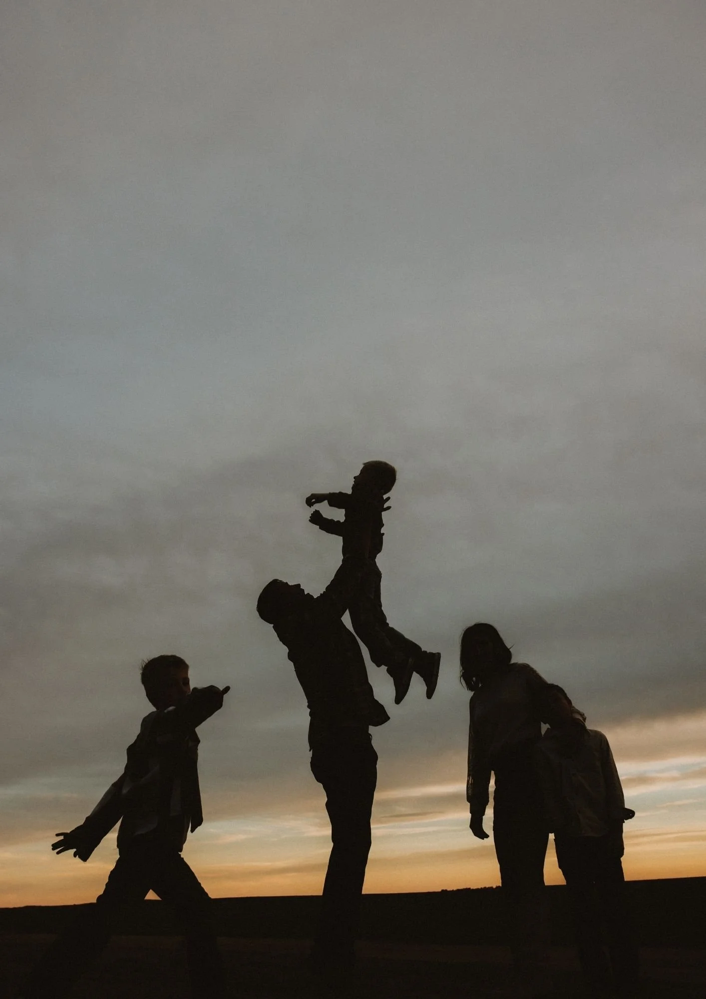Silhouettes of four people outside at sunset, with one person holding a child up in the air, against a cloudy sky.