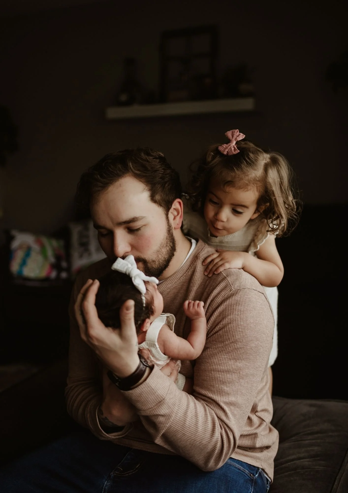 A man holding a newborn baby, with a young girl leaning over his shoulder. The man is kissing the baby's forehead, and the girl is looking at the baby affectionately.