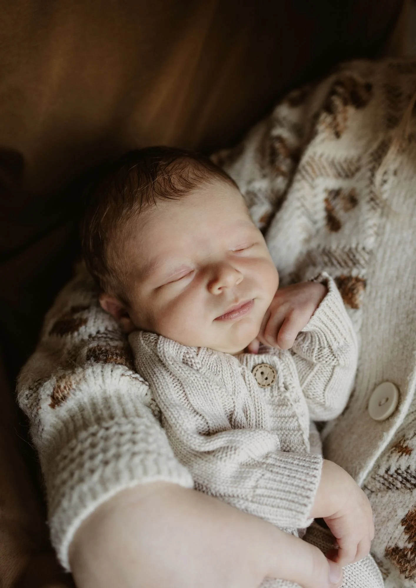 A sleeping baby wearing a cream-colored, knit sweater, resting on a patterned blanket.