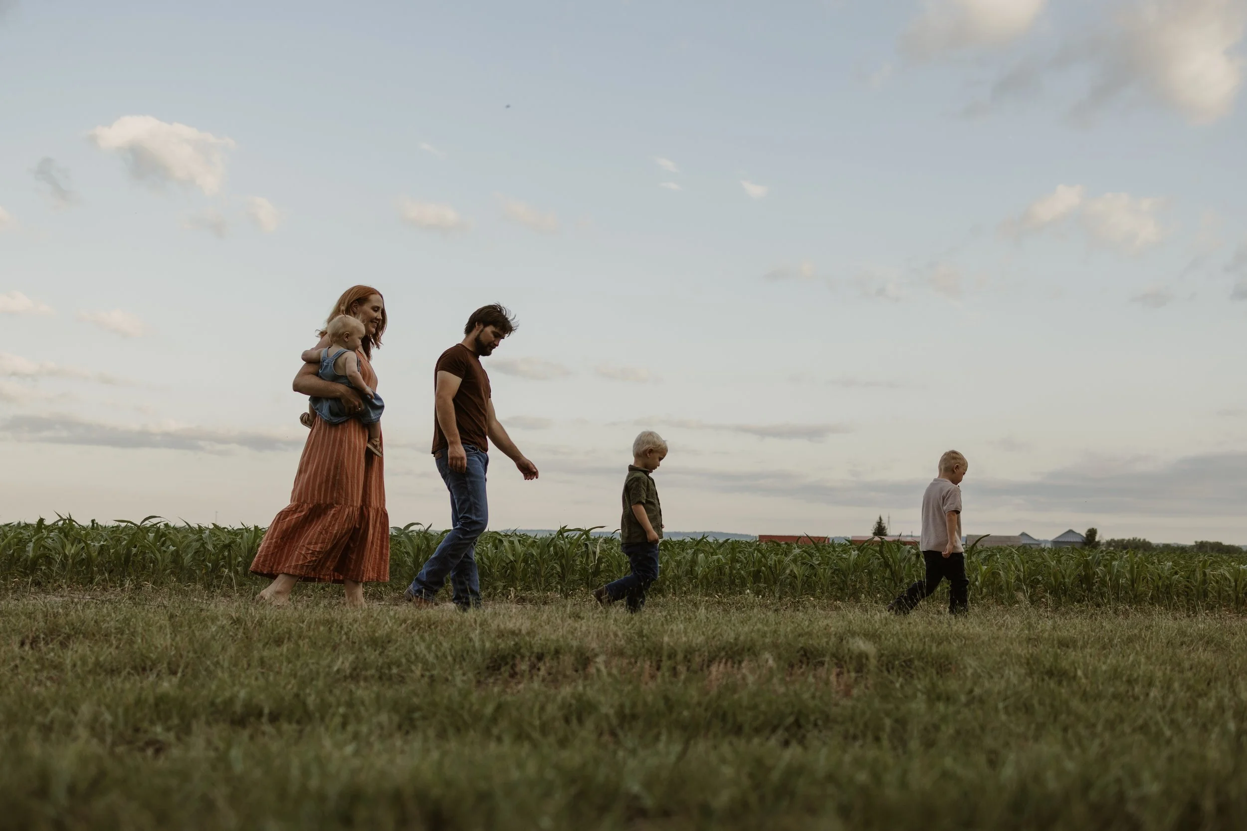 A family of five walking in a field near a corn farm during sunset, with the mother holding a young child and the father walking beside two other children.