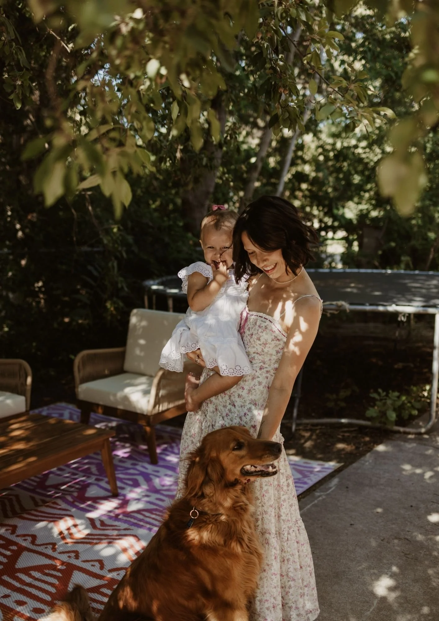 A woman holding a young girl outside in a shaded area with trees, a garden, outdoor furniture, and a golden retriever dog nearby, all smiling and enjoying a sunny day.