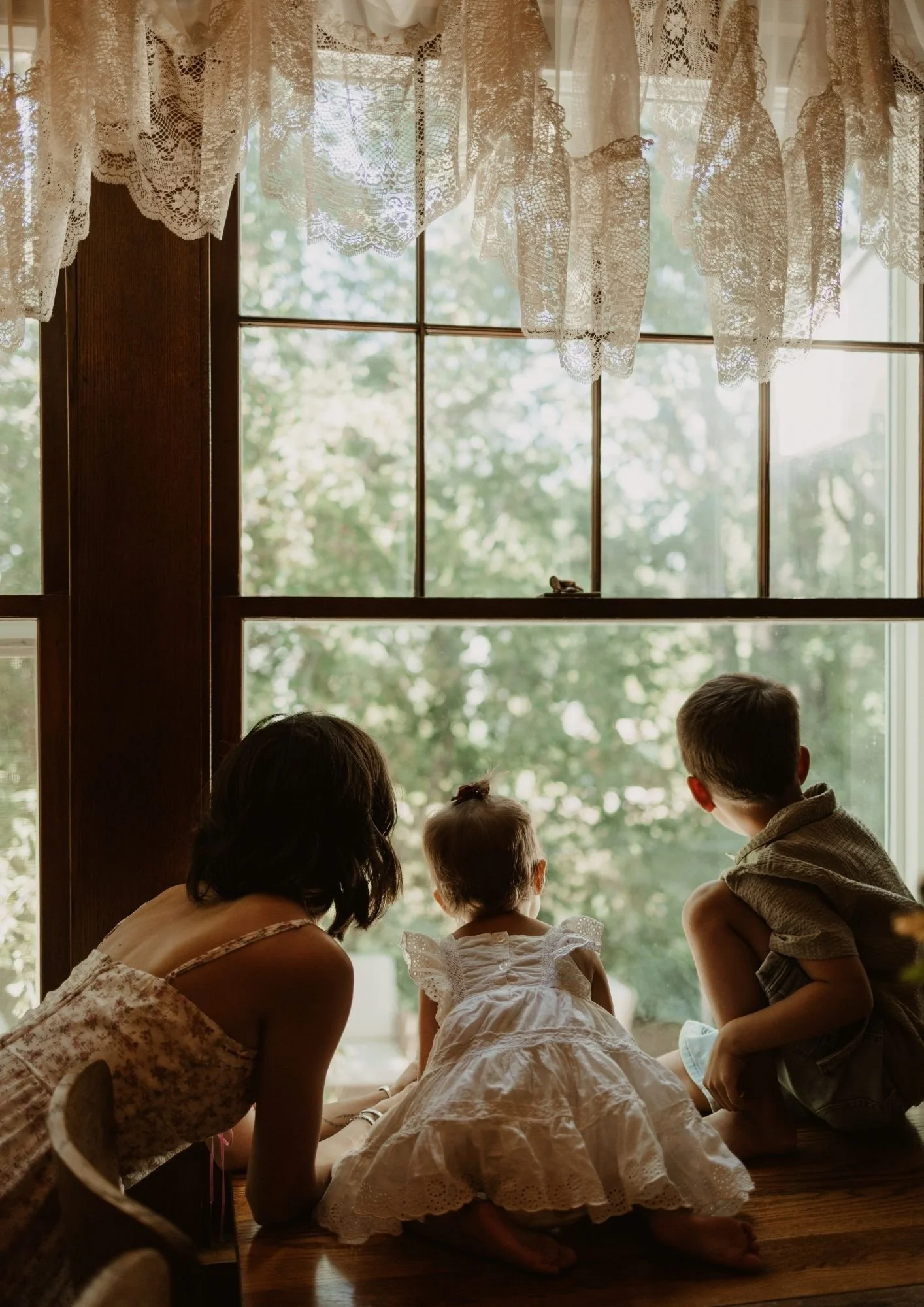 A woman and two young children sitting on a wooden windowsill, looking outside through a window with lace curtains, with greenery visible outside.