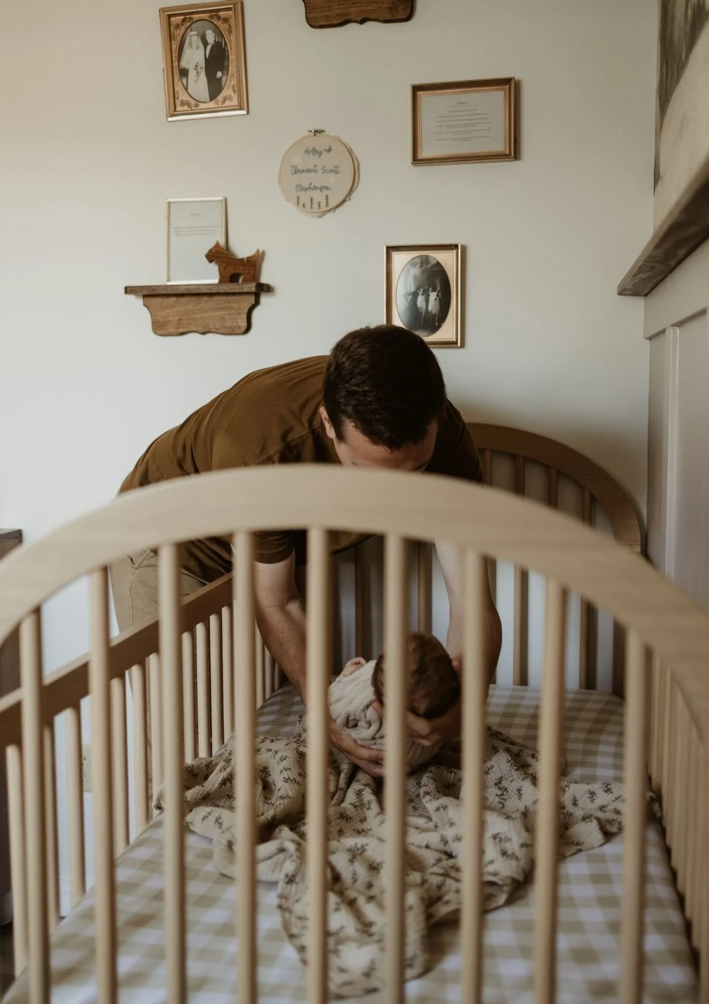A man leaning over a crib helping a baby inside, with framed photos and decorations on the wall behind them.