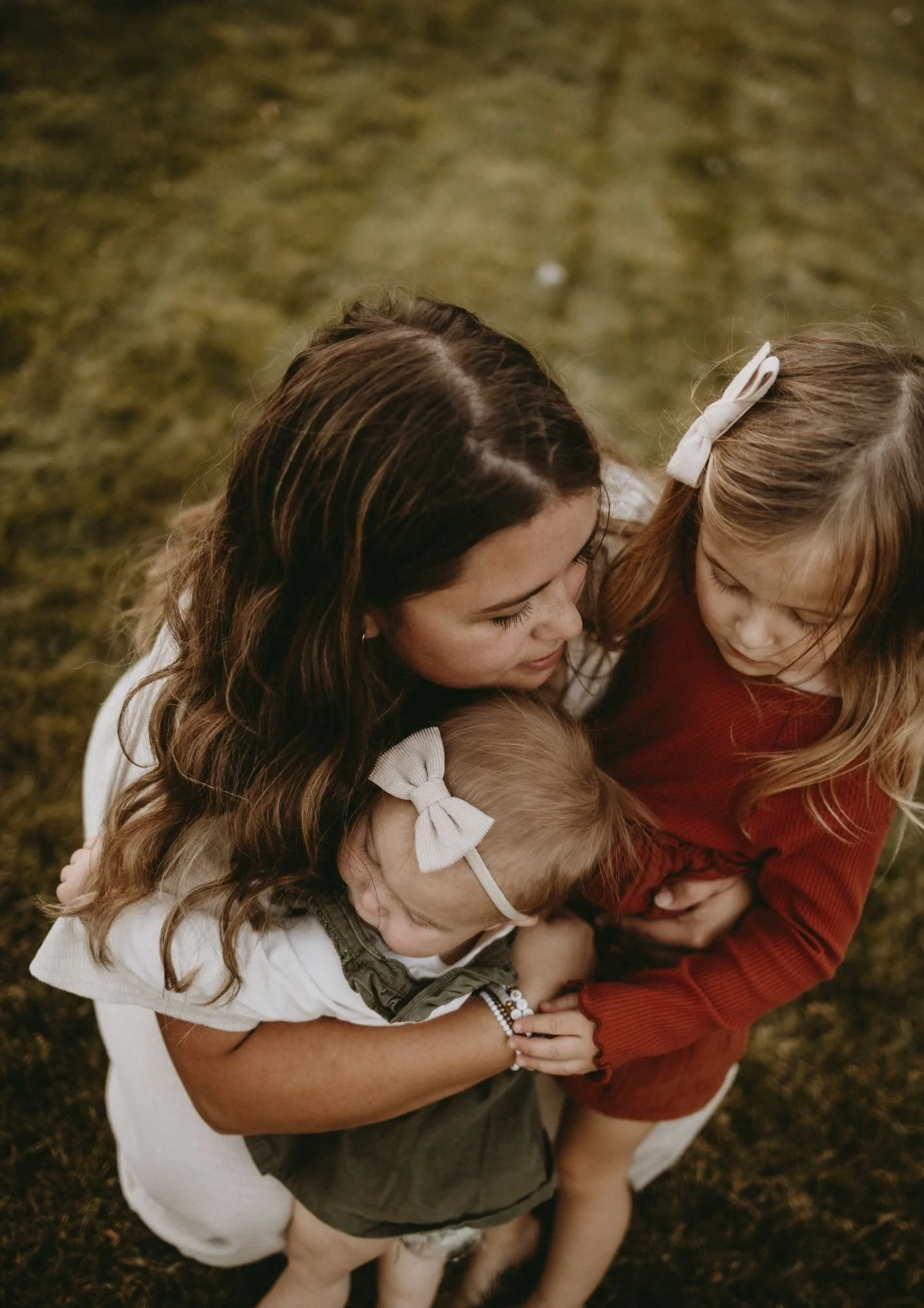 A woman with long brown hair hugging two young girls, one with curly blonde hair and a large white bow, and the other with straight reddish-brown hair and a white bow, outdoors on grass.