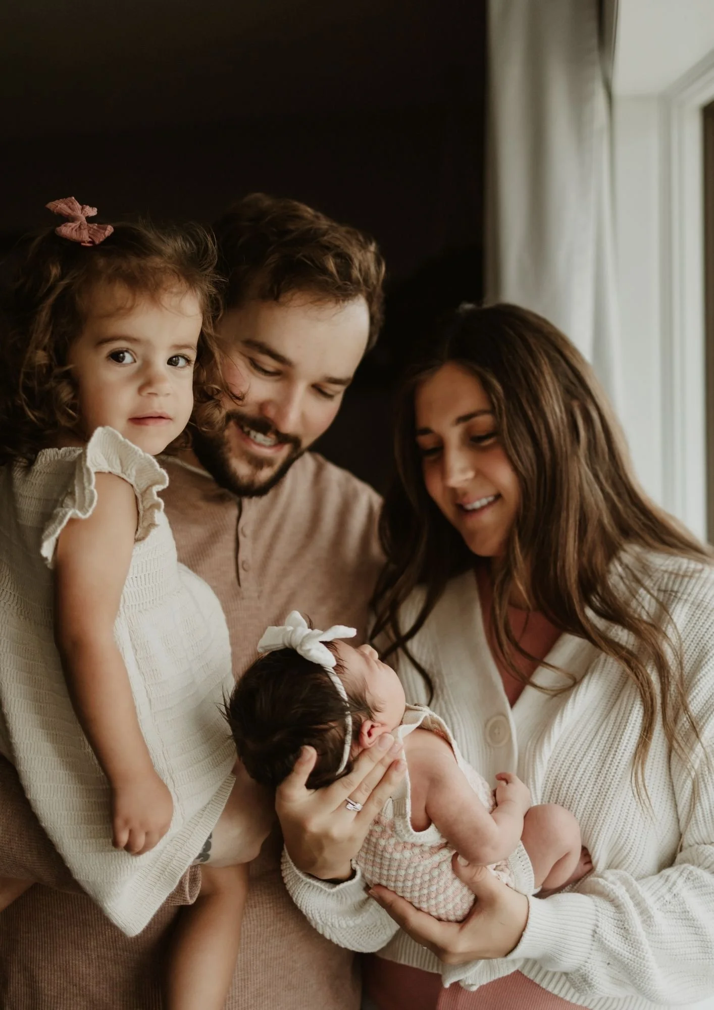 Family of four with two young children, one being a newborn, gathered indoors near a window, with warm lighting and smiling faces.