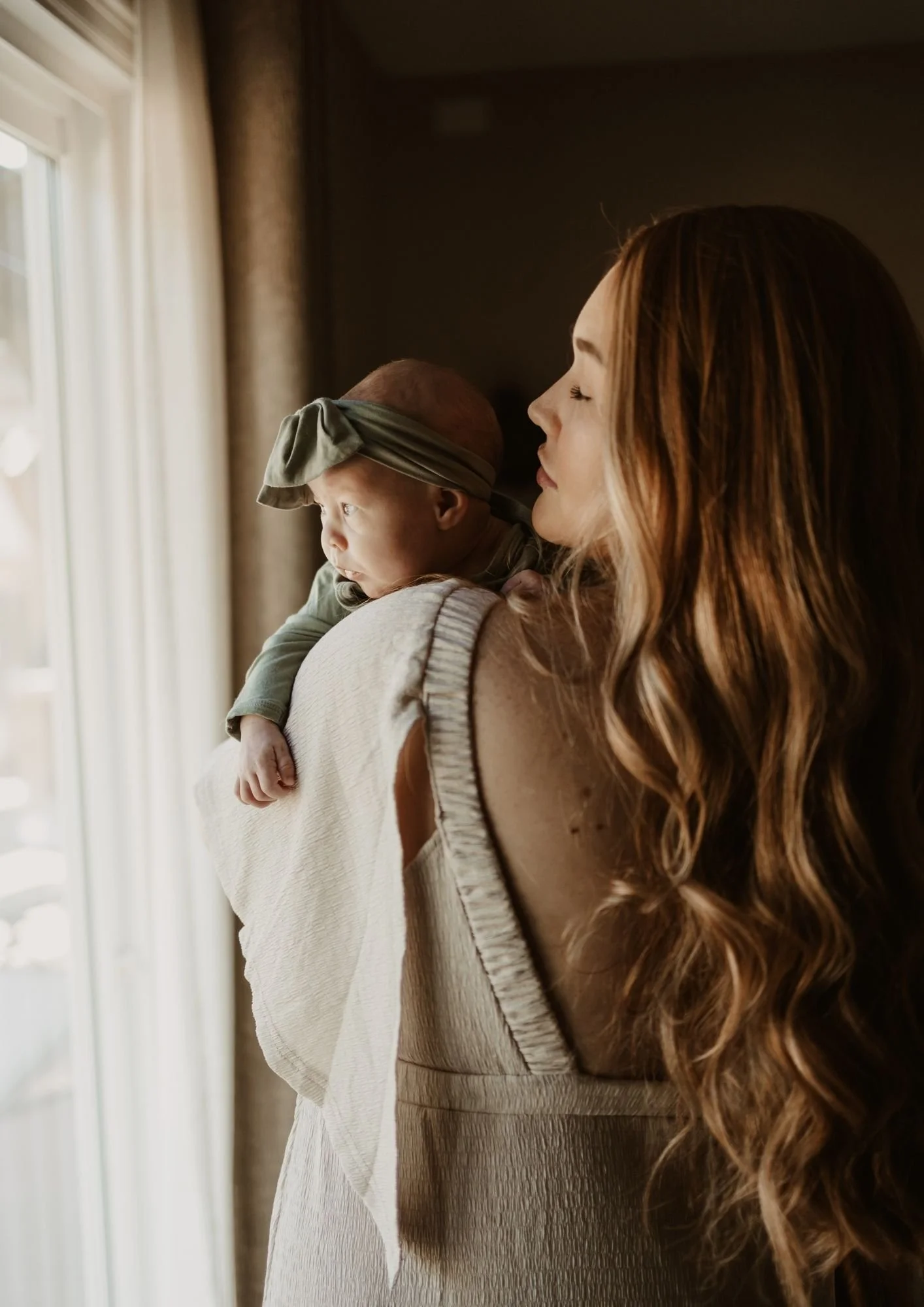 A woman with long wavy hair holding a baby boy with a green headband near a window, looking outside.