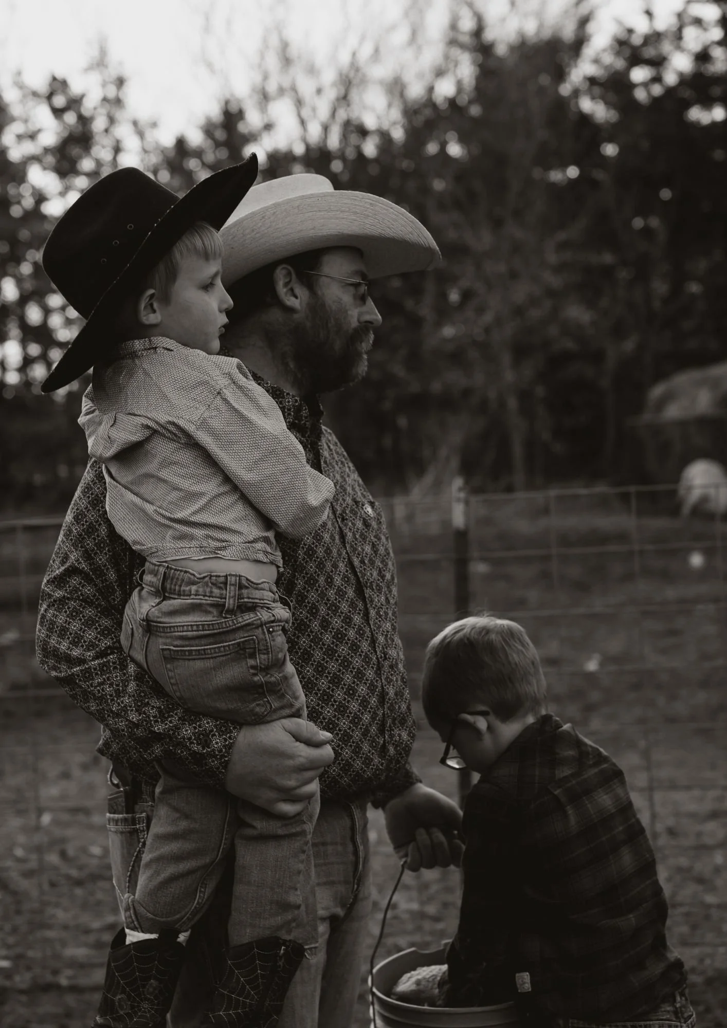 A man wearing a large cowboy hat holds a young boy with a cowboy hat on his lap. Another boy wearing glasses kneels in front of them, looking into a bucket. All are outdoors in a rural setting with trees in the background.