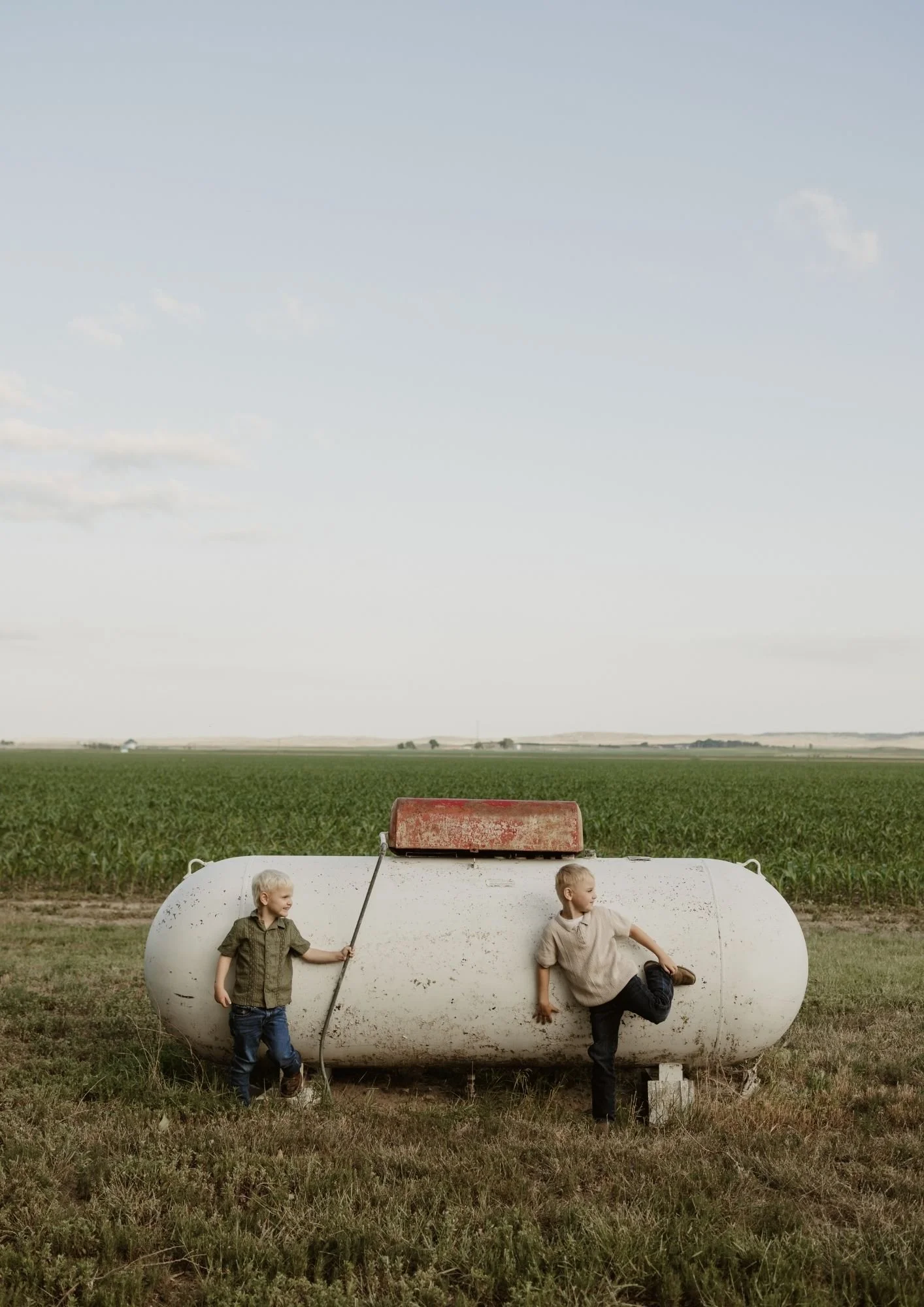 Two young boys with blonde hair playing near a large, old, white propane tank in a rural field, one boy standing and holding a stick, the other leaning against the tank with one leg raised, under a cloudy sky.