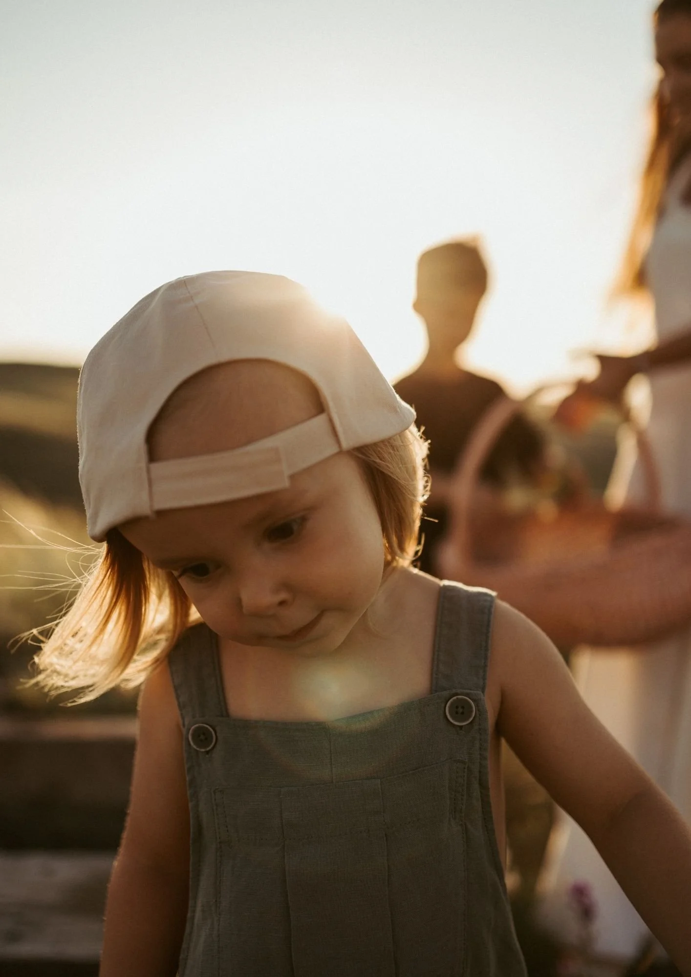 A young child with red hair wearing a beige baseball cap backward and a gray sleeveless jumpsuit, looking down in front of a sunlit background with blurred figures.