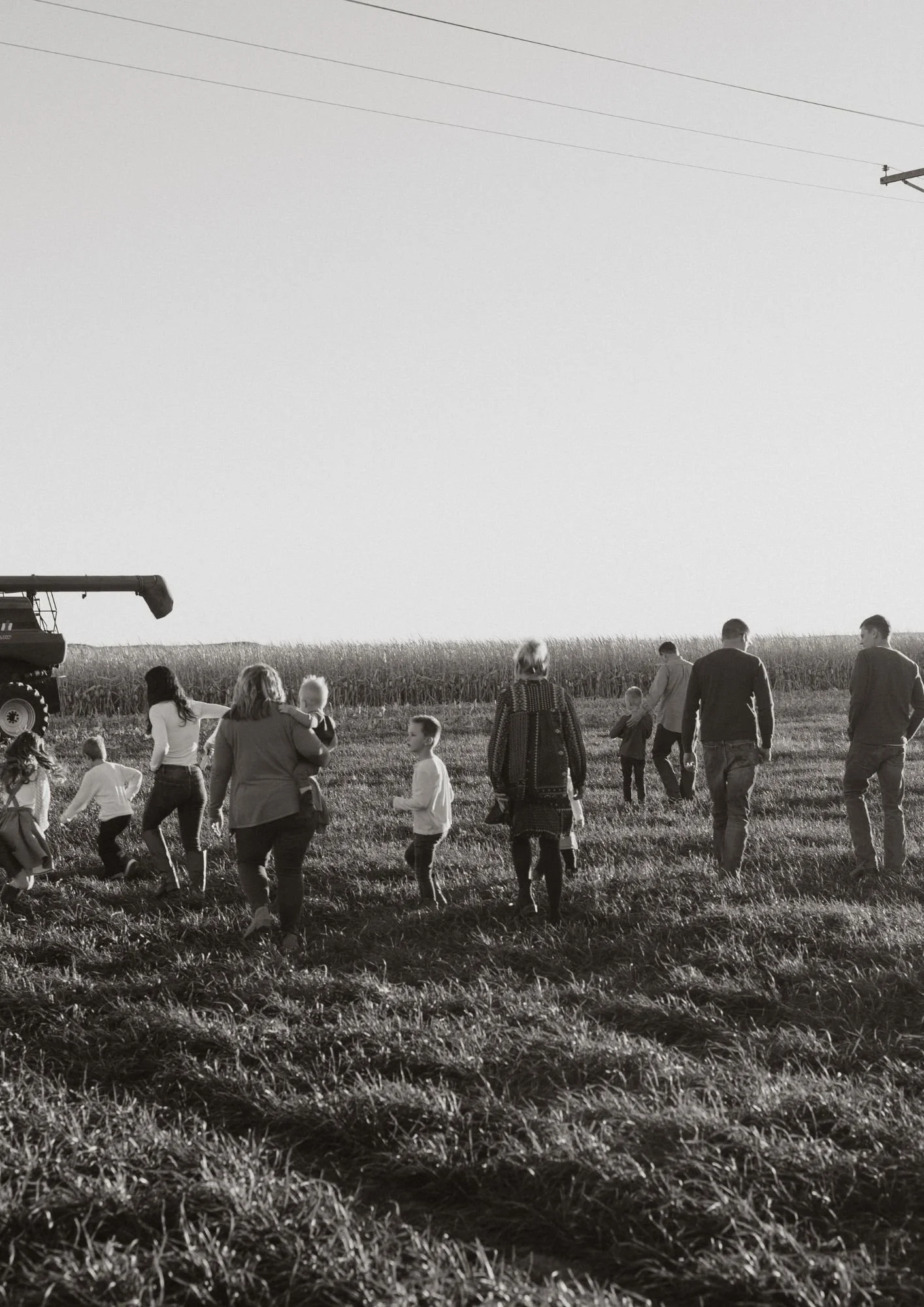 A black and white photo of a group of people, including children and adults, walking and playing in a grassy field near a farm, with a tractor on the left and power lines overhead.