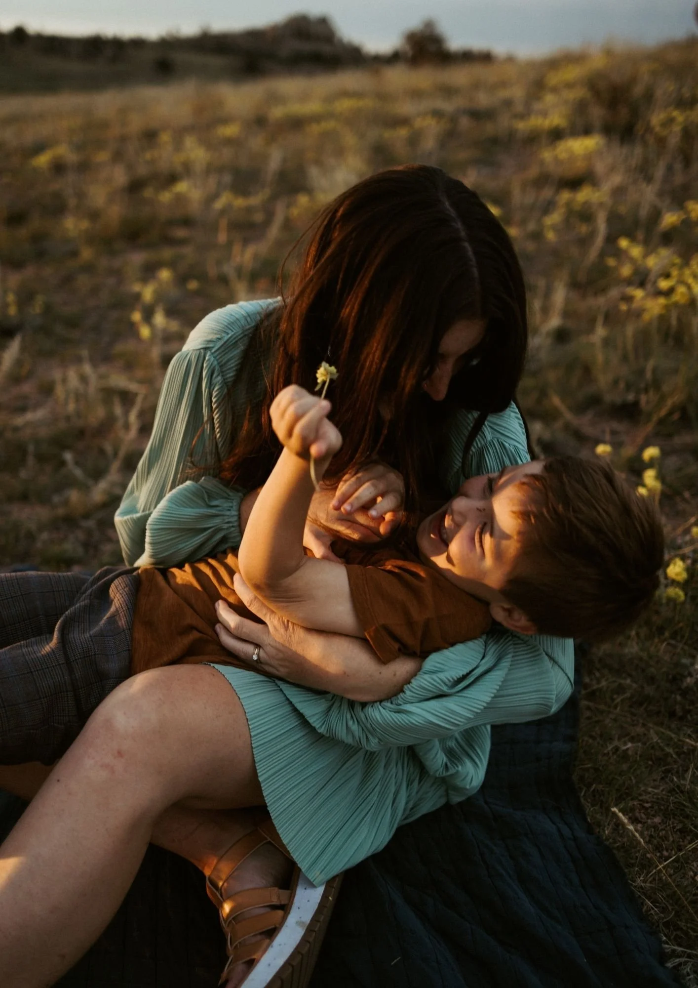 A woman and a boy playing and cuddling in a field during sunset, with the woman holding a small flower and smiling at the boy.