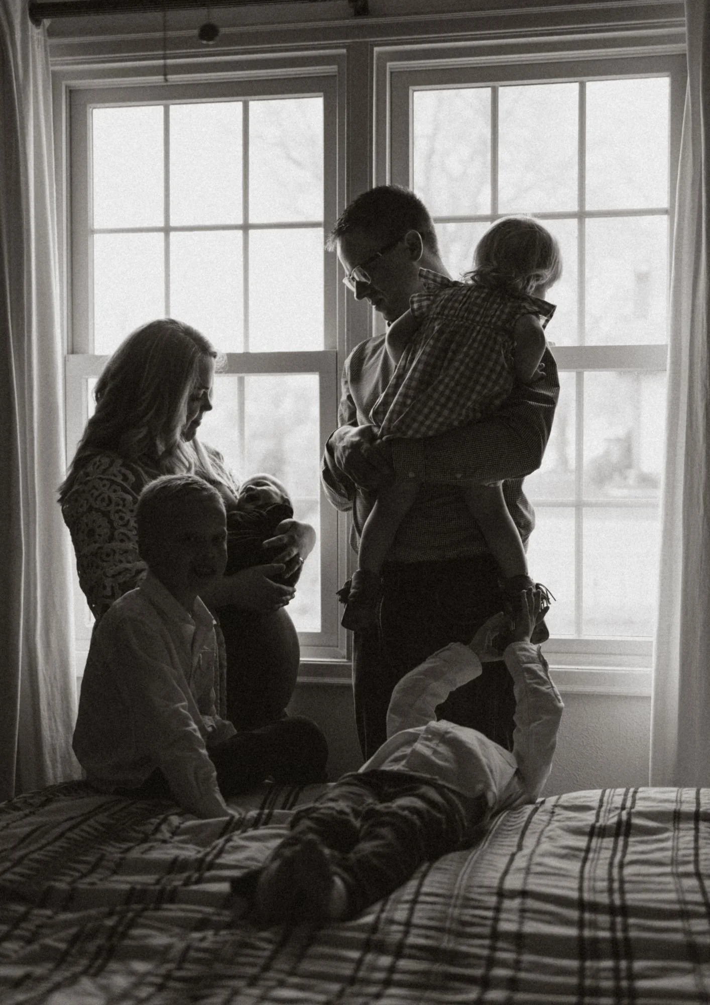 A black and white photo of a family scene with five children and two adults in front of a window. One child is laying on a bed in the foreground, while another is seated on the bed. The adults are holding babies, and the scene is backlit by the windo