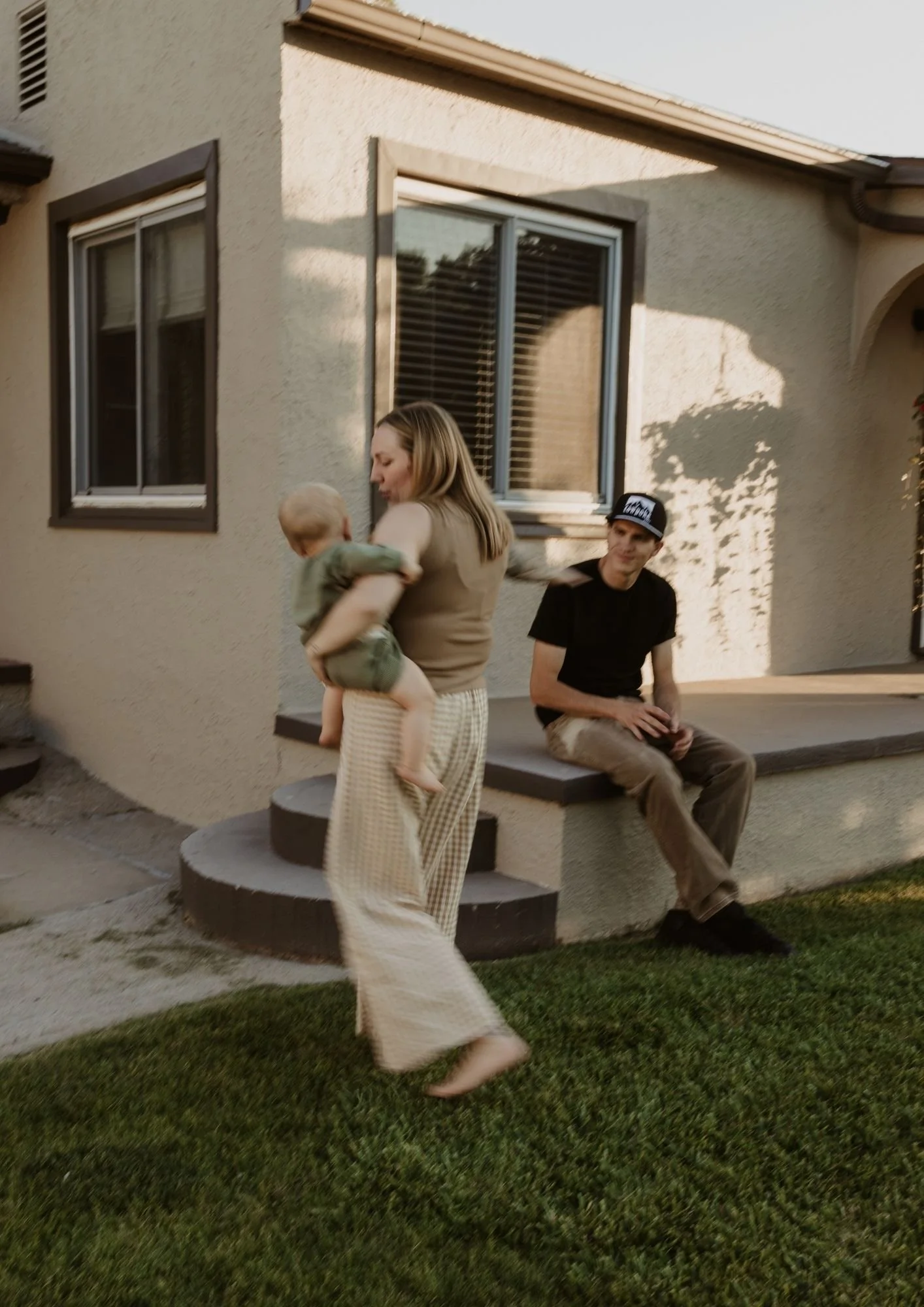 A woman holding a toddler in her arms, standing on a step outside a house, with a man sitting on the ledge nearby, looking at her, during what appears to be late afternoon or early evening.