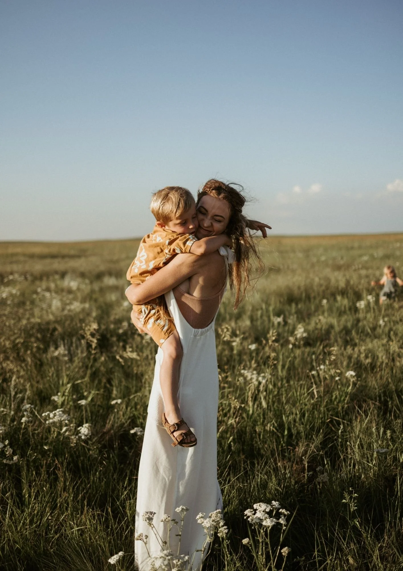 A woman holding a young boy in a grassy field with wildflowers, both smiling and hugging, with a clear blue sky in the background.