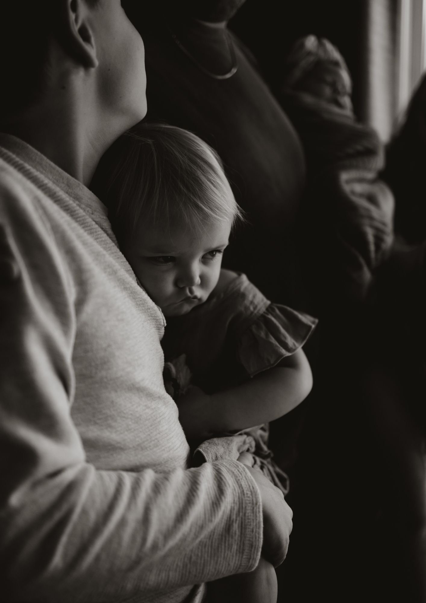 A young child is held by an adult, sitting in a dark room near a window with light coming in. The child appears to be pouting or upset, with a serious expression. The photo is in black and white.