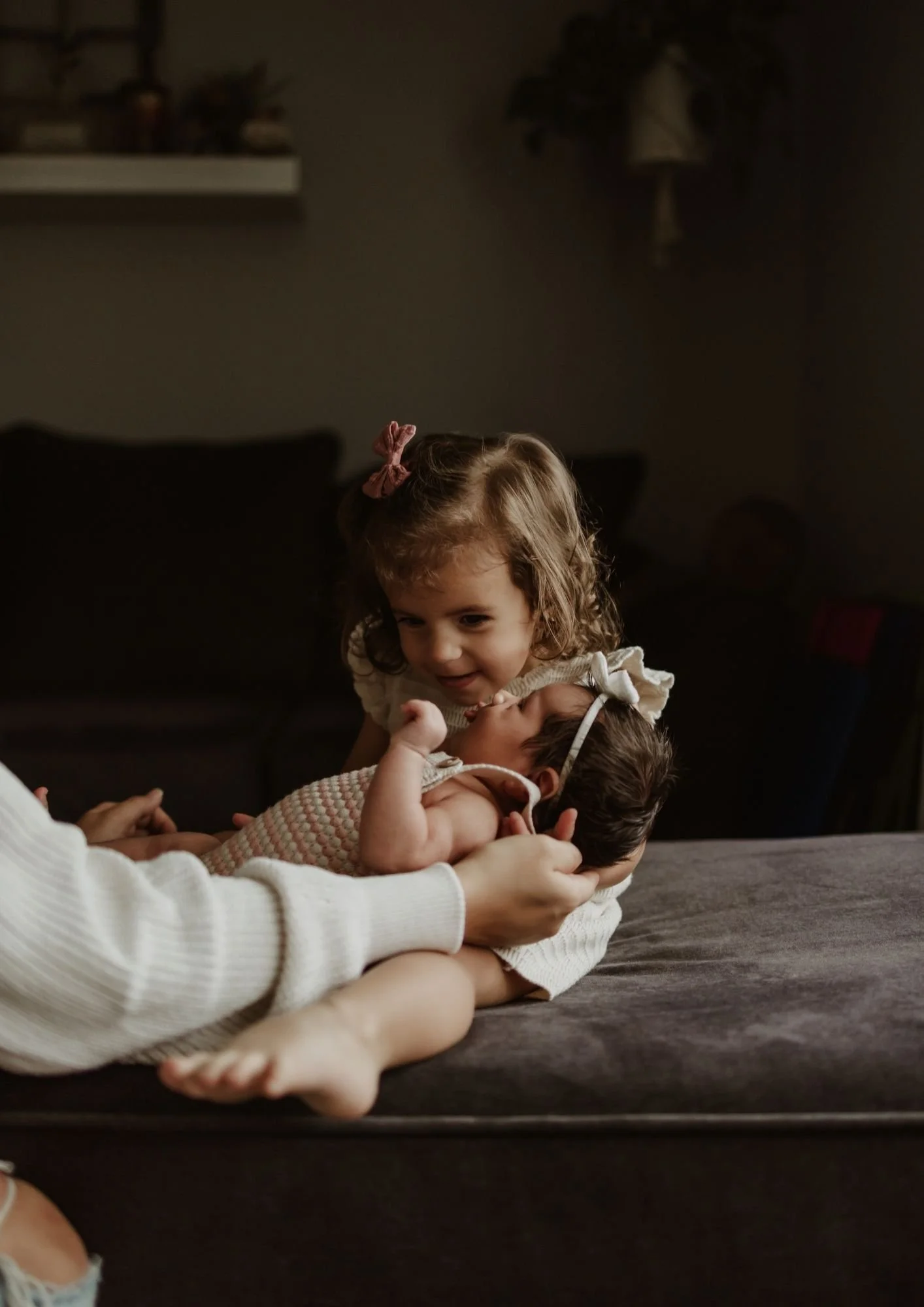 Two young children, one with curly hair and a pink bow, are playing with a newborn baby on a bed. The older girl is smiling and leaning over, while the baby is lying down, wearing a knitted outfit and a headband. An adult's hands gently hold the baby
