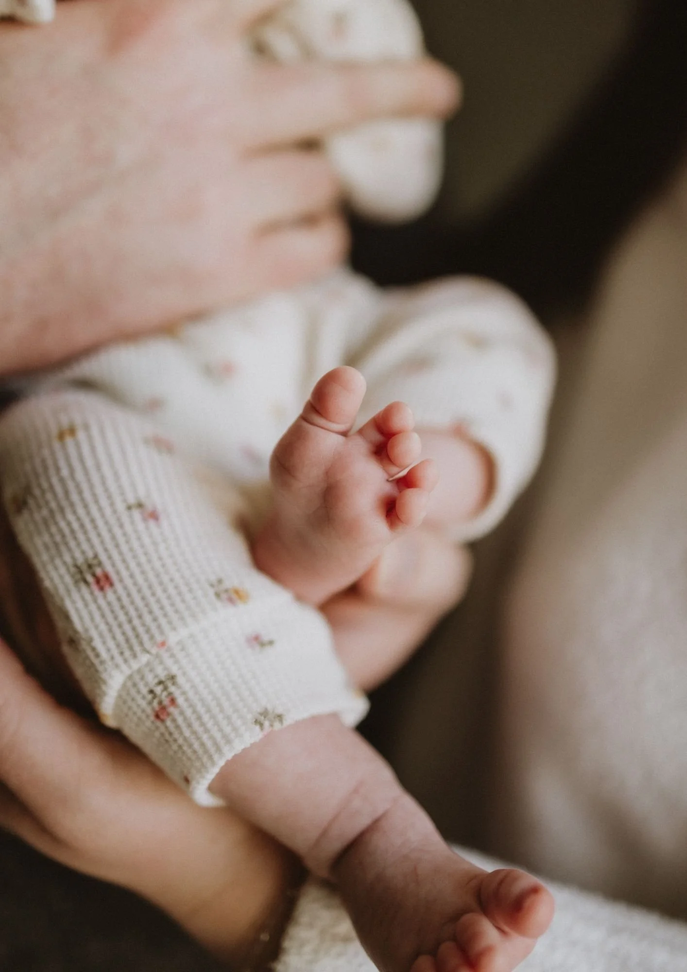 Close-up of a newborn baby's tiny hand clasped in an adult's hand, with the baby's face slightly out of focus in the background.