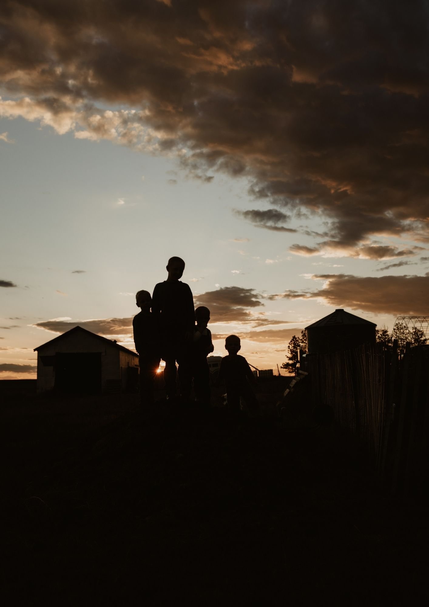 Silhouettes of four children standing outdoors during sunset, with farm buildings and cloudy sky in the background.