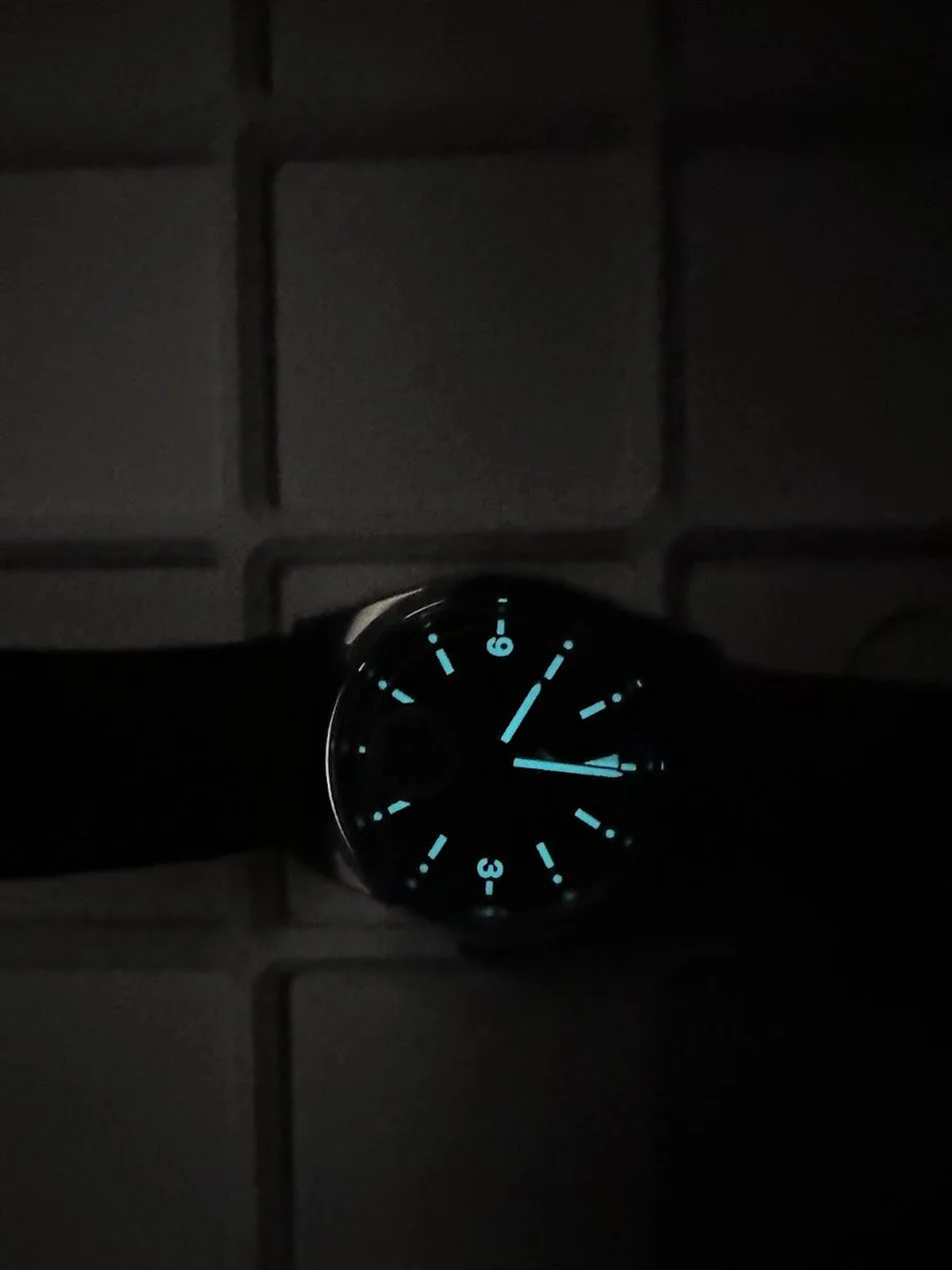 A wristwatch with a black strap, showing a dark face with glowing blue hour markers and hands, lying on a tiled surface in low light.