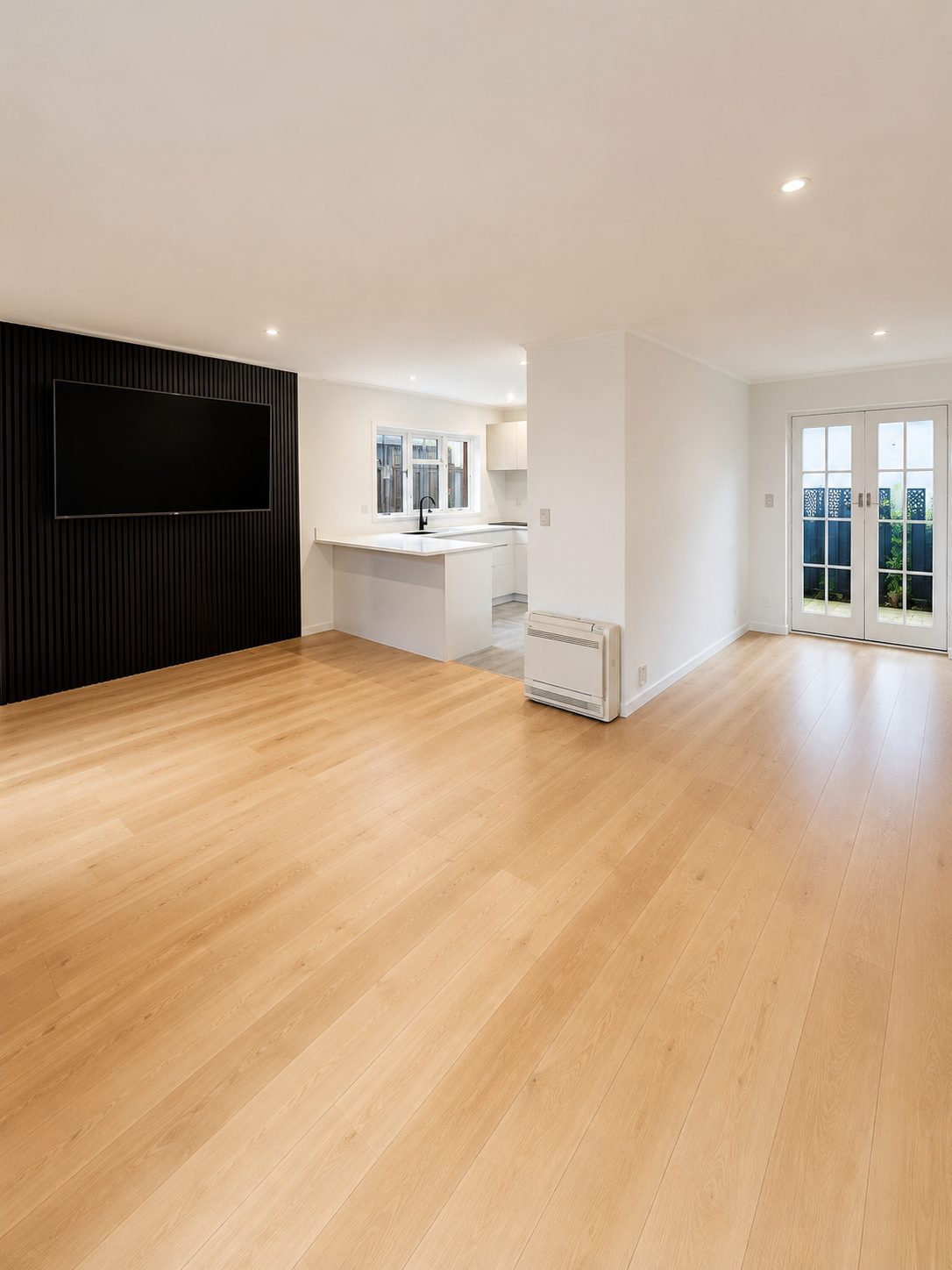 Empty living room with hardwood floors, white walls, a black accent wall with a mounted flat-screen TV, a small heater on the wall, and doors leading outside.