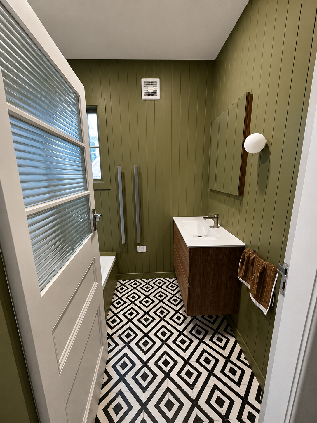 Small bathroom with green paneled walls, black and white patterned floor, wall-mounted wooden cabinet with white sink, brown towel hanging on the side, small window, white door with glass panels, and modern light fixture.