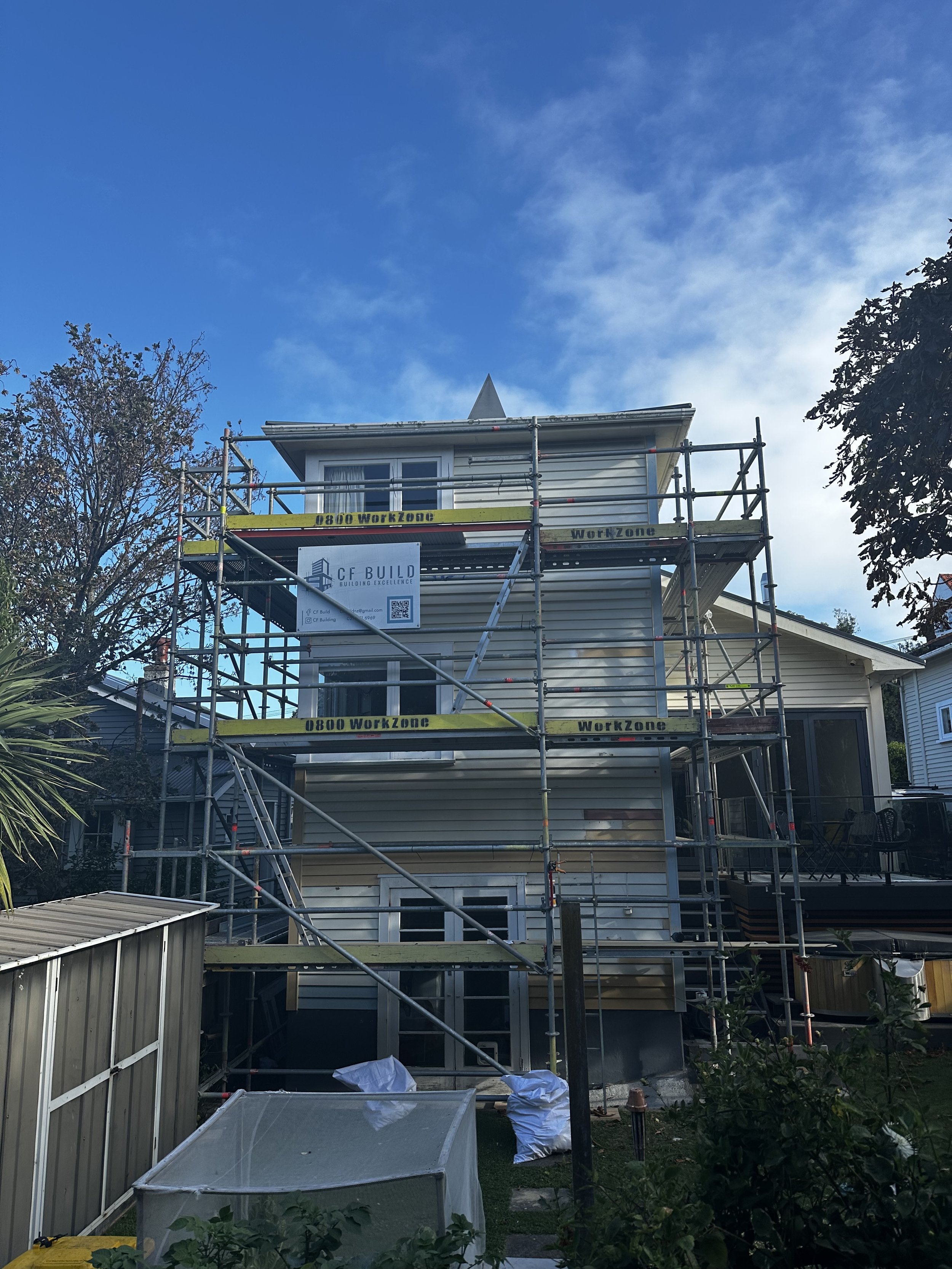 A house under construction with scaffolding around it, featuring a sign that reads "CF Build" and "0800 WorkZone." The house has beige siding, several windows, and a small deck or balcony with outdoor furniture. The sky is partly cloudy with some blue visible.
