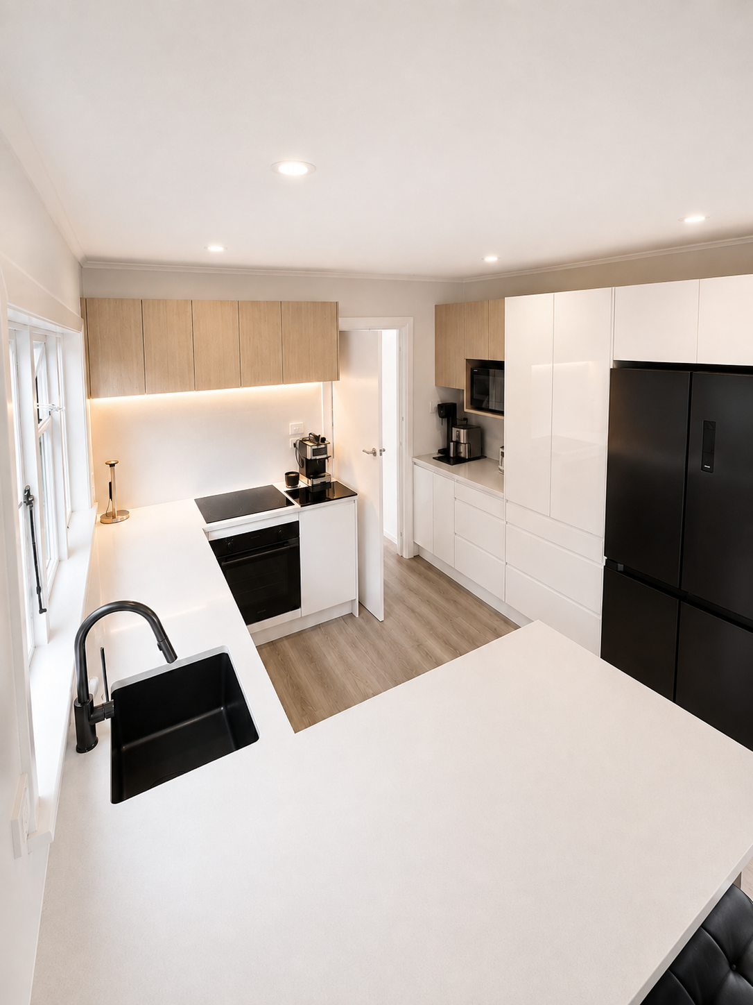 Modern kitchen with white countertops, black appliances, and a breakfast bar with black chairs. There are cabinets, a coffee maker, and a microwave, with a small window on the left and a doorway at the back.