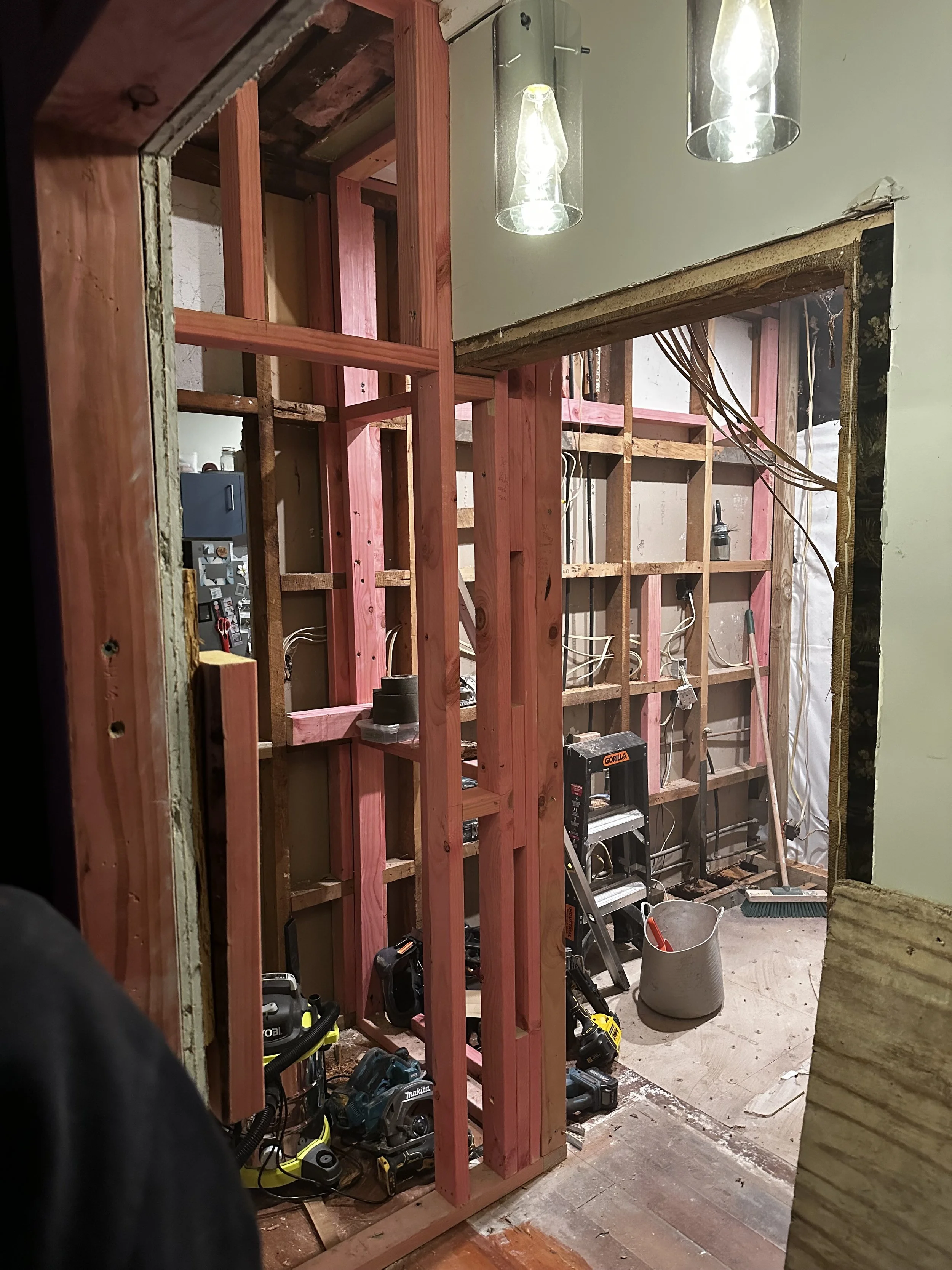 Construction site with wooden framing, electrical wiring, tools like drills, and work lights hanging from the ceiling.