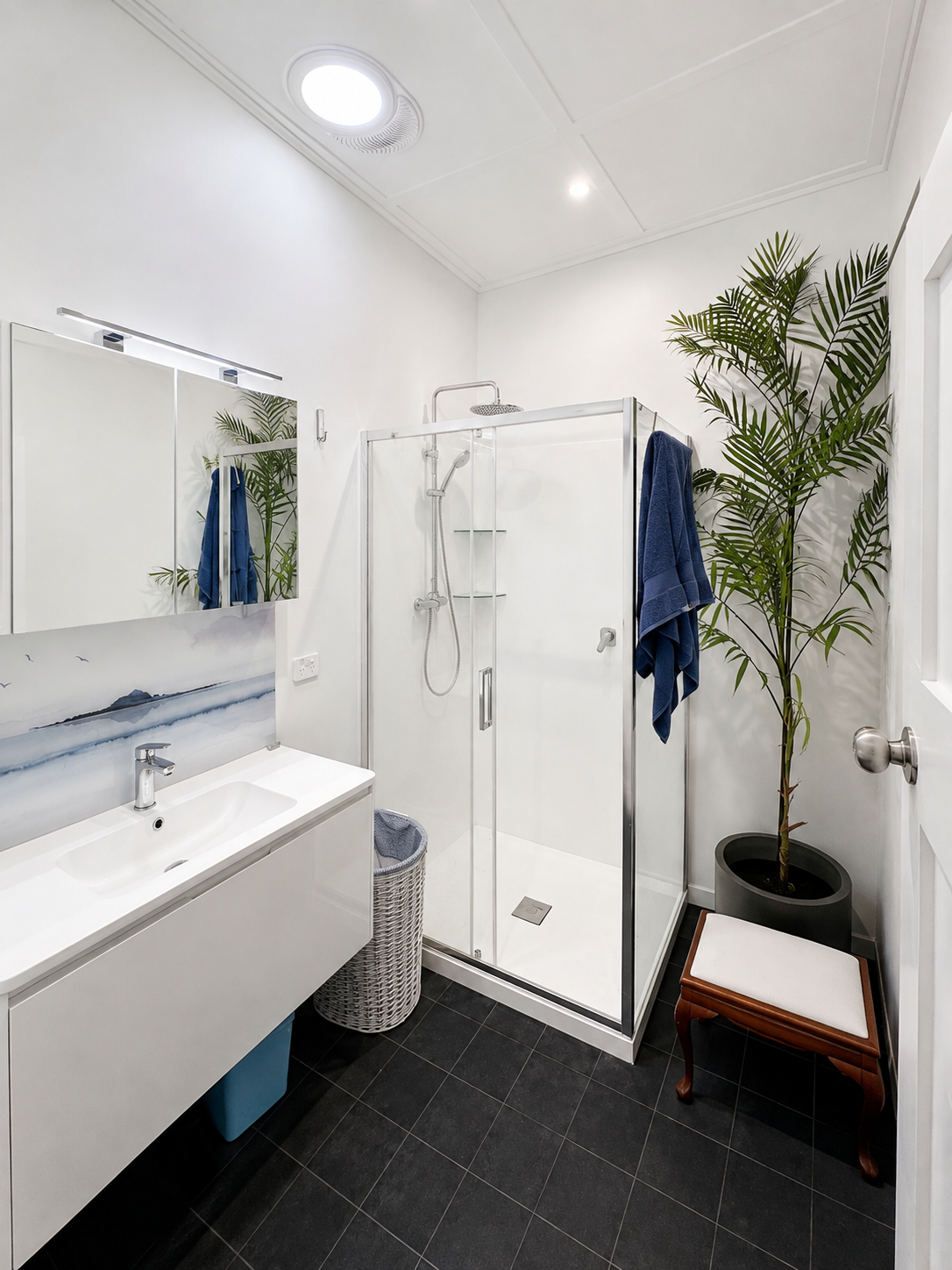 A modern bathroom featuring a glass shower enclosure, a white sink with a marble backsplash, a mirror, a laundry hamper, a large potted plant, a wooden stool, and blue towels hanging.