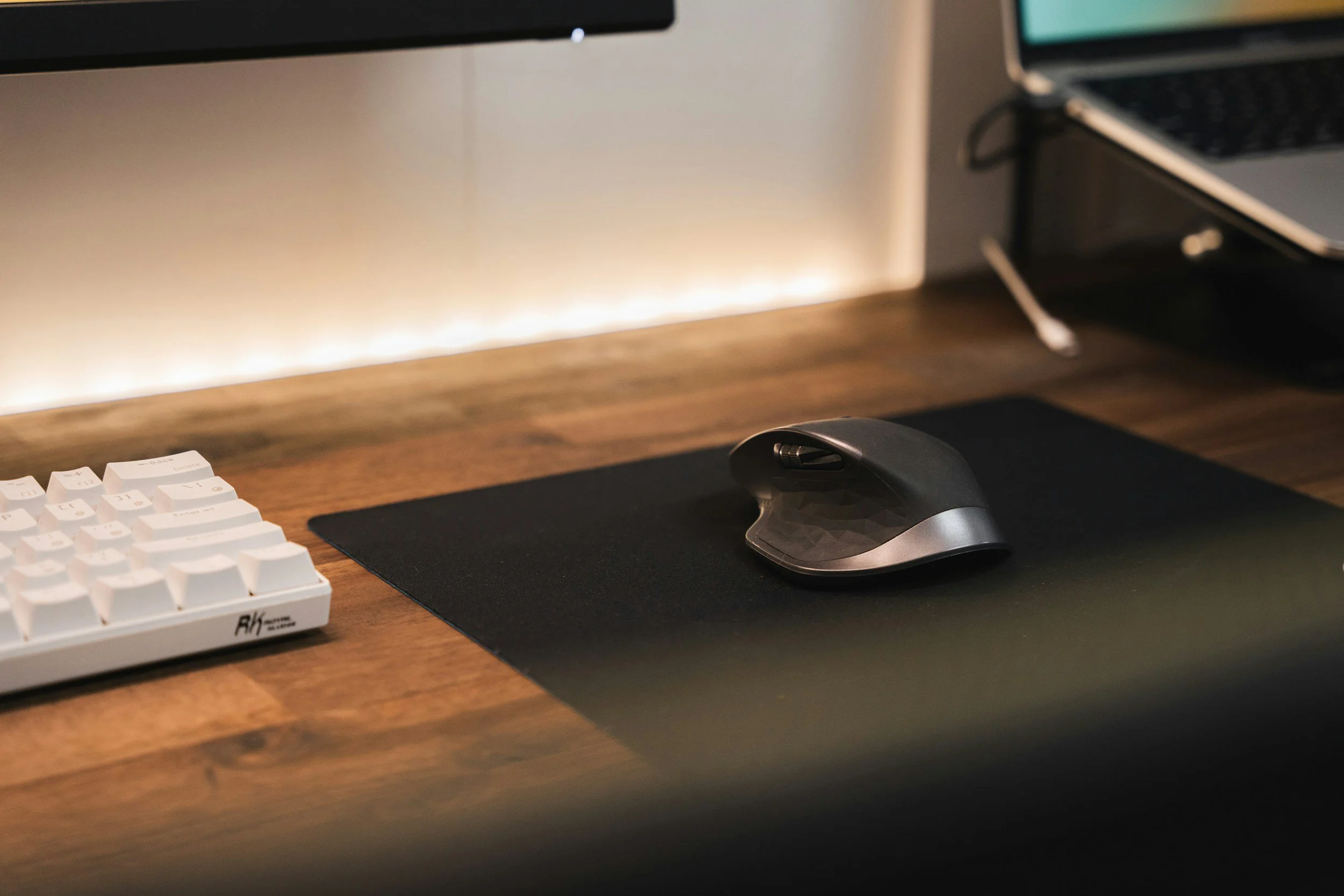 A modern workspace with a wooden desk, a black mouse on a mouse pad, a white keyboard, and a laptop with a screen, illuminated by soft LED strip lighting.