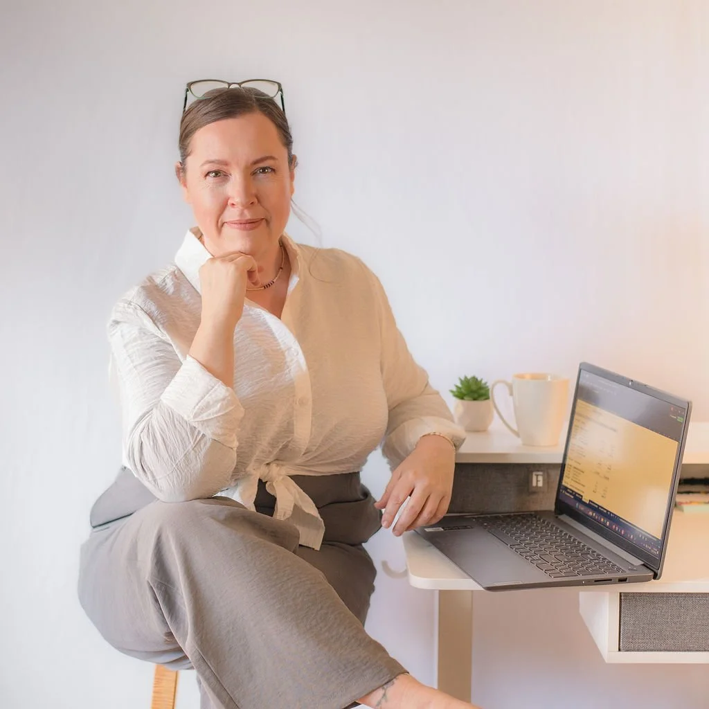 Valerie, owner of VLK Bookkeeping, sitting on a chair at a desk with a laptop, a mug, and a small potted plant, looking at the camera.