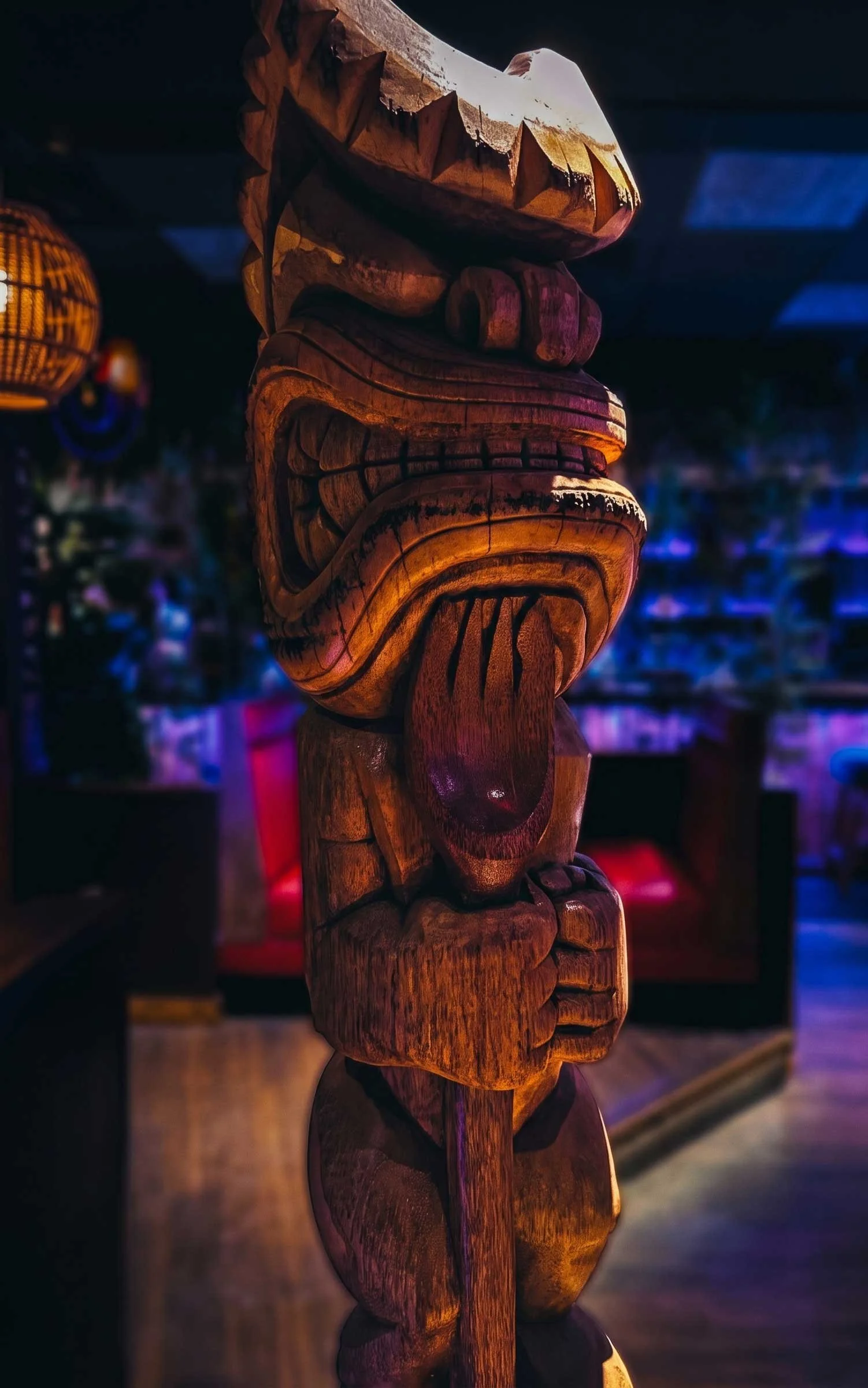 Close-up of a hand-carved wooden tiki statue with a fierce, toothy grin and outstretched tongue, lit in warm amber lantern light against the dark, moody blue and purple neon glow of a tiki bar interior.