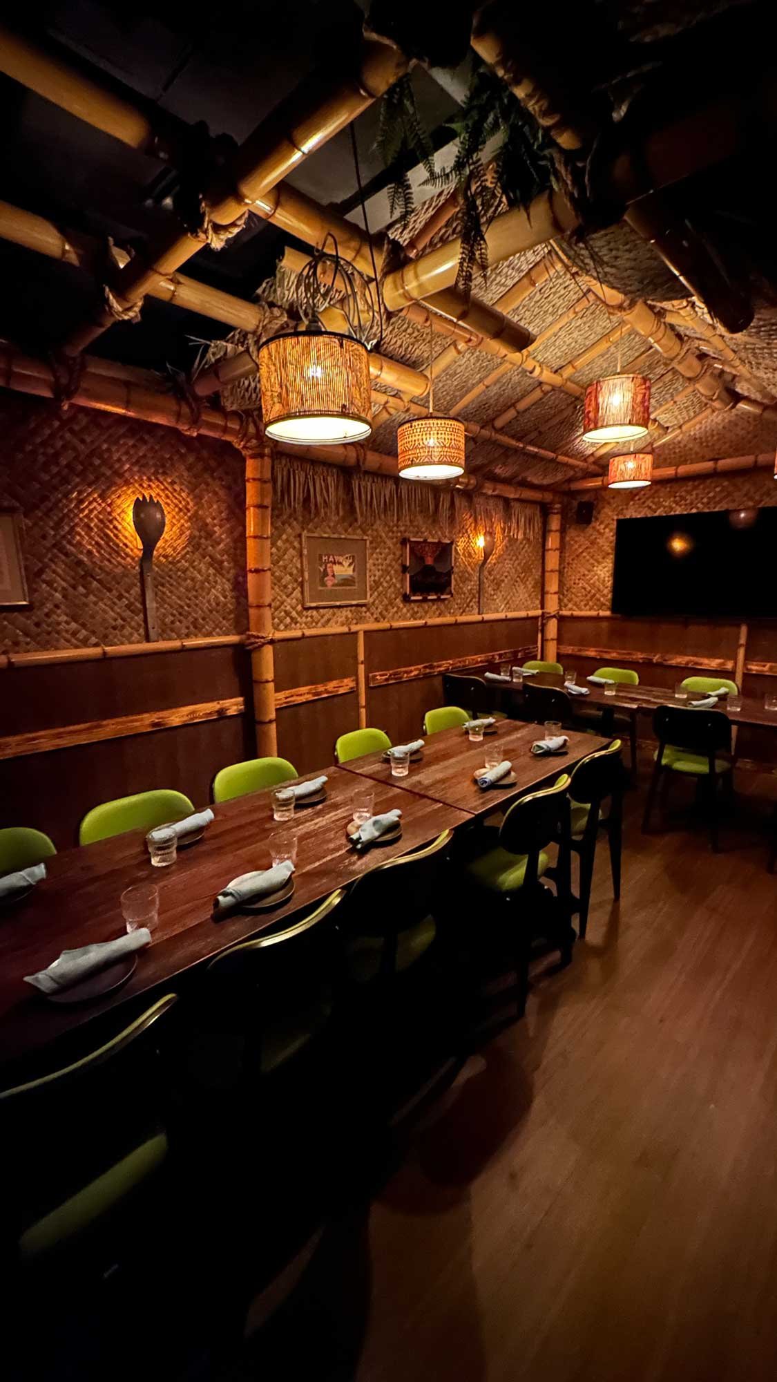 Private event dining space at Spork Island Trading Company featuring long tables, tropical bamboo décor, warm pendant lighting and a wall-mounted TV.