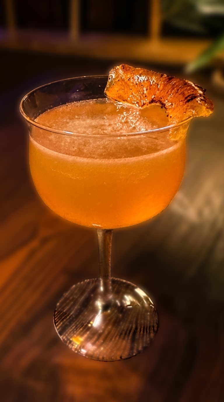 A frothy orange cocktail in a coupe glass garnished with a charred pineapple wedge on the rim, set on a wood bar table with a blurred tiki backdrop of bamboo and greenery