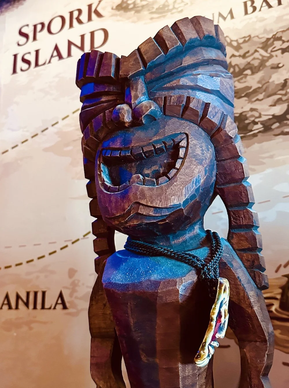 Hand-carved wooden tiki statue with a toothy grin and cowrie-shell necklace, lit in blue and purple light against a vintage 'Spork Island' map backdrop."