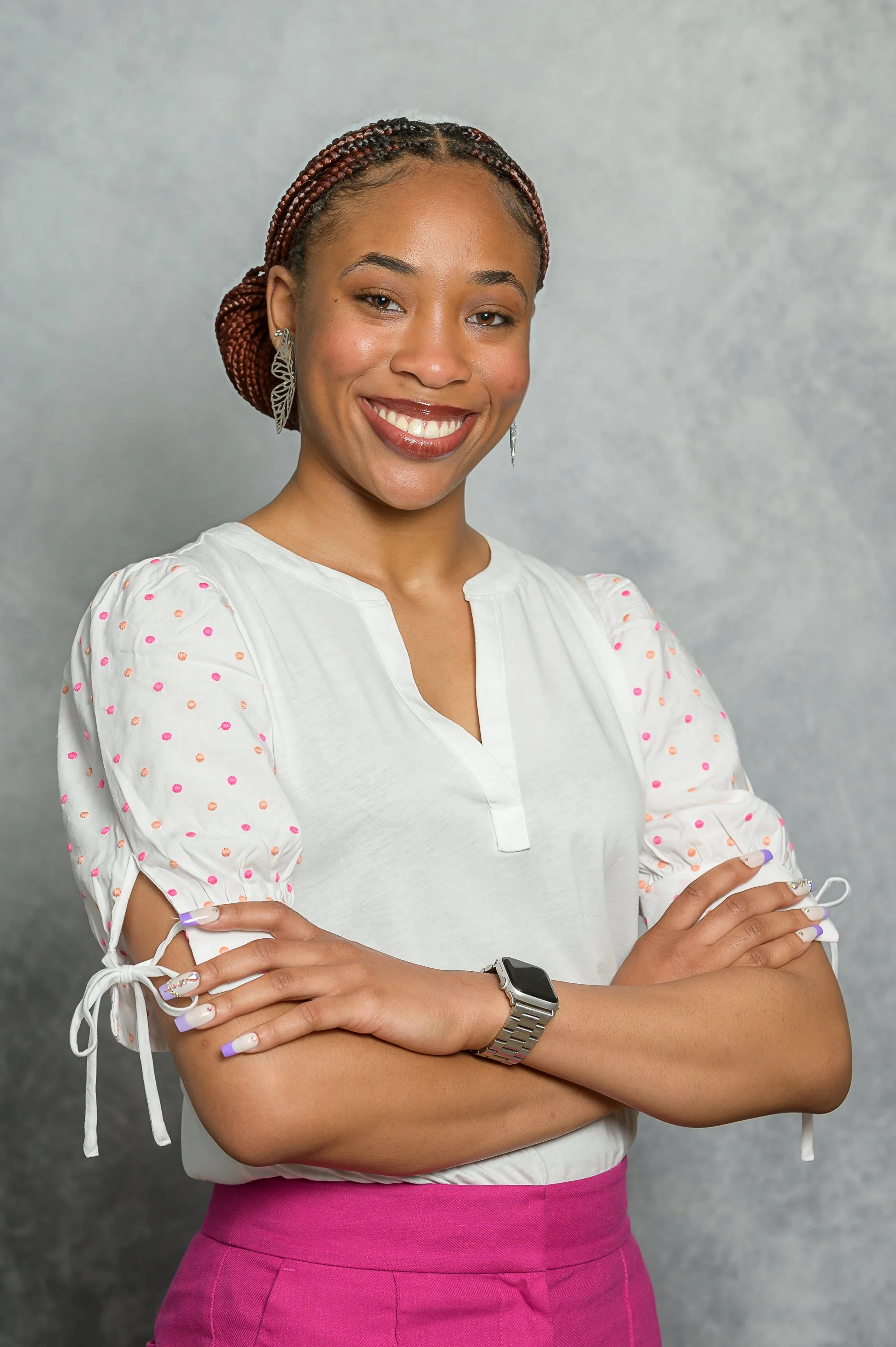 Portrait of a young woman with braided hair, wearing a white blouse with pink polka dots on the sleeves and pink skirt, smiling with arms crossed in front of a gray background.