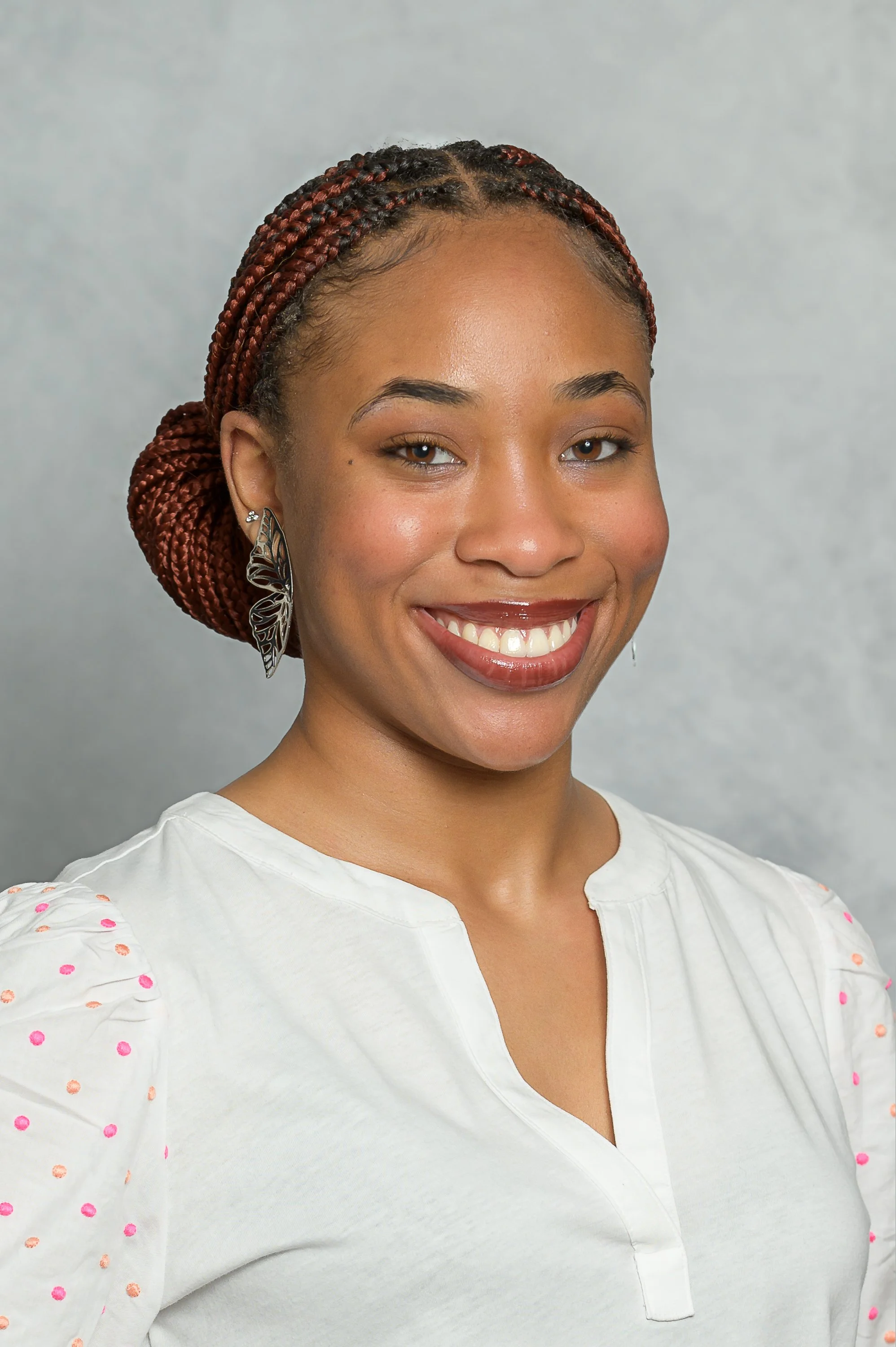 A woman with braided reddish-brown hair styled in an updo, wearing a white blouse with pink polka dots on the shoulders, large butterfly-shaped earrings, and smiling at the camera against a neutral gray background.