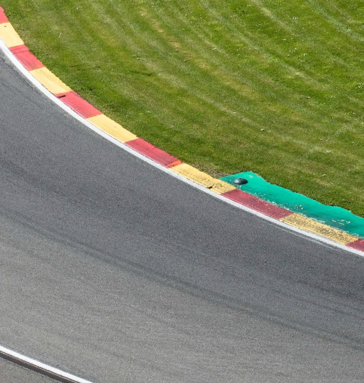 Close-up of a racing track curb with red and yellow painted sections, next to a grassy area, with a portion of the asphalt racing surface visible.