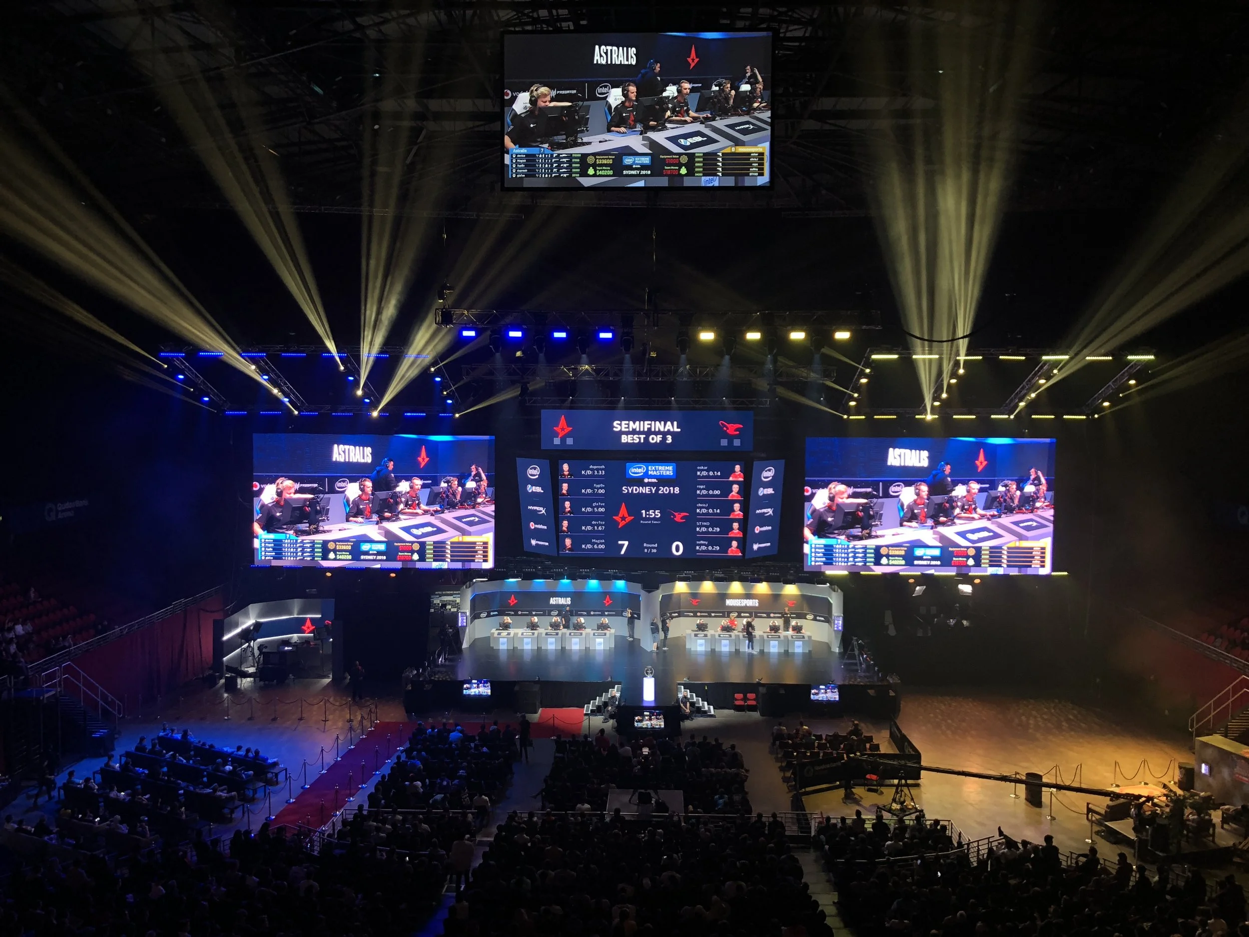 An esports tournament stage with large screens displaying a game between Astralis and other teams, showing players seated at computer stations, with bright yellow and blue lighting and an audience watching from below.