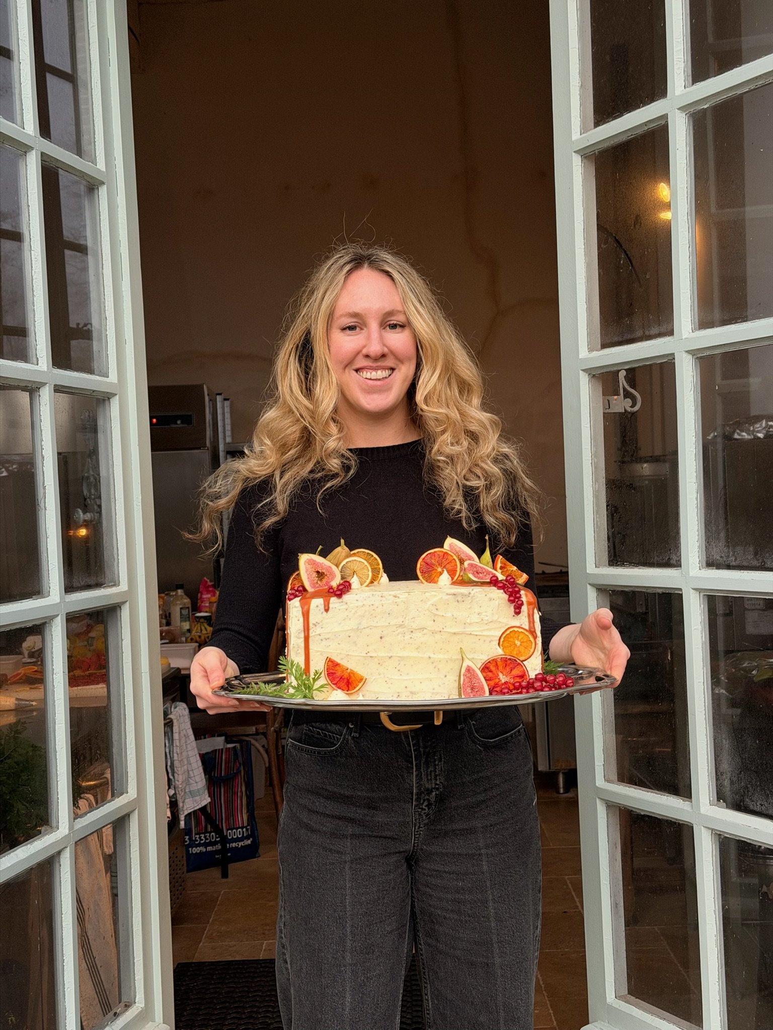 A woman standing in a doorway holding a decorated birthday cake with fruit toppings.