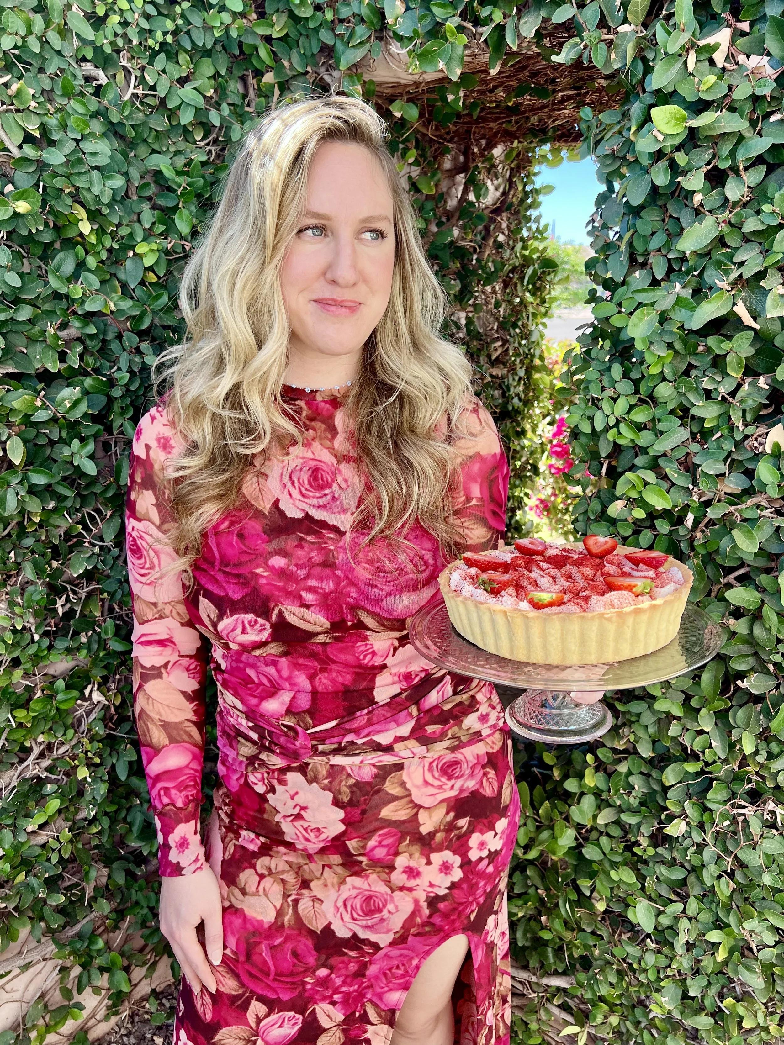 A woman in a pink floral dress holding a strawberry tart stand outdoors in front of green shrubbery.